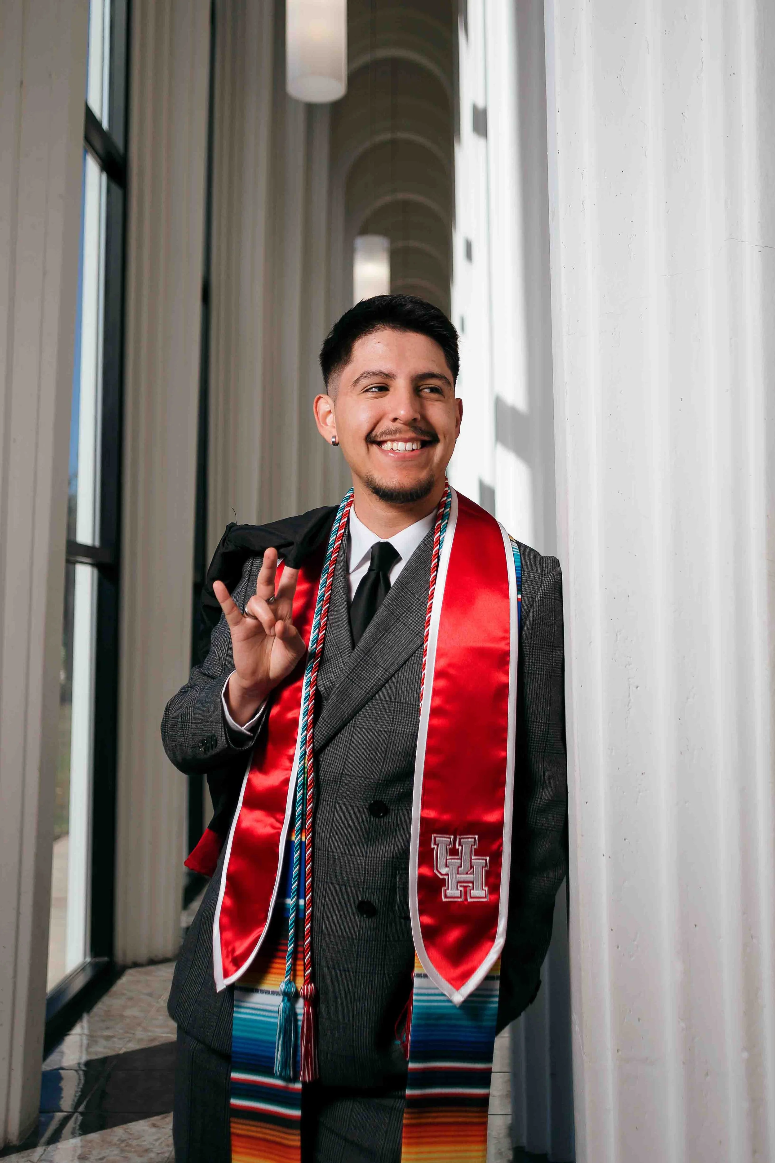 University of Houston graduation portrait by Lev’s Photography featuring a young man in a suit and tie wearing a red stole and honor cords, smiling while holding his graduation gown over his shoulder inside a modern campus corridor