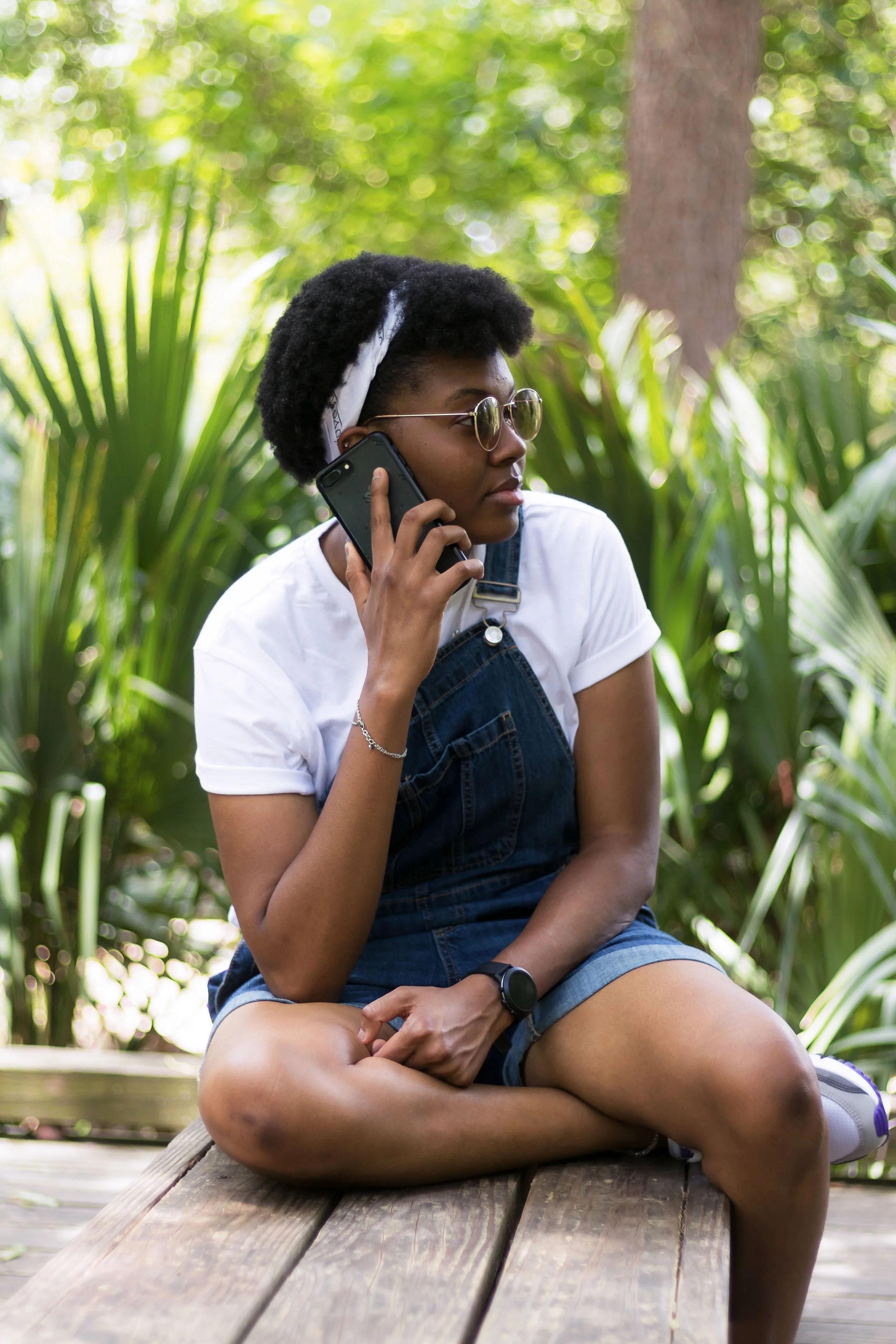 A woman sitting on a wooden bench outdoors, talking on a cellphone, surrounded by lush green plants and trees.