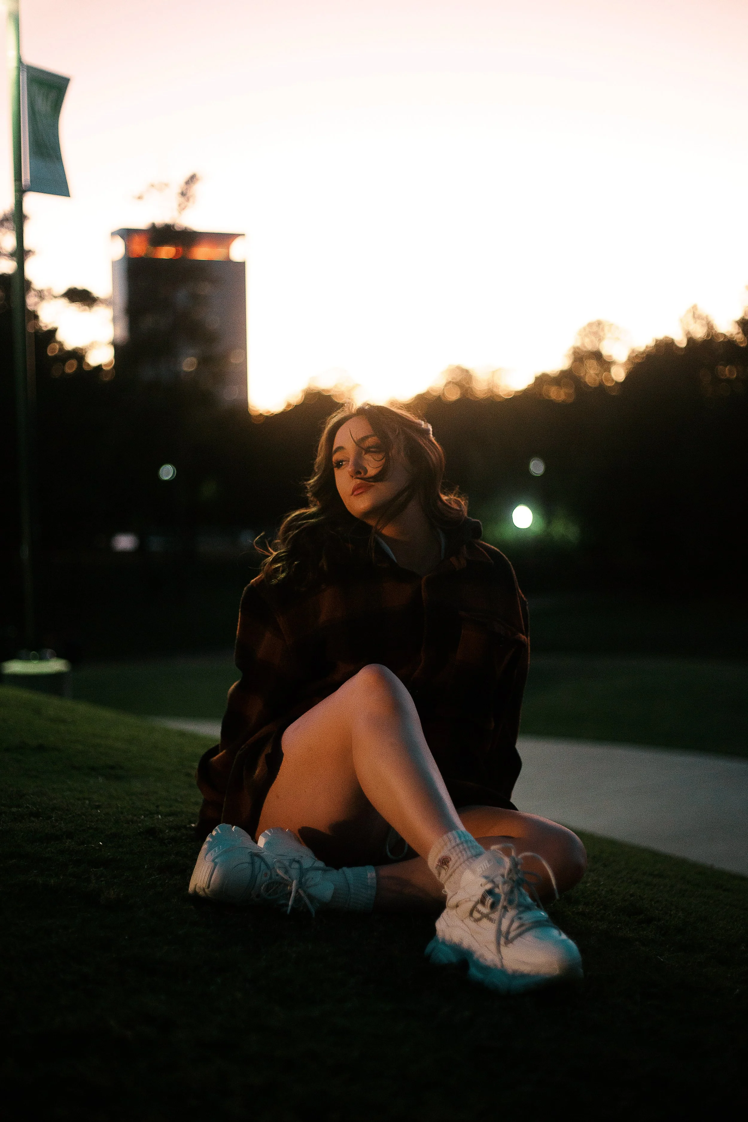 A young woman sitting on grass at sunset, wearing sneakers, shorts, and a jacket, with trees and buildings in the background.