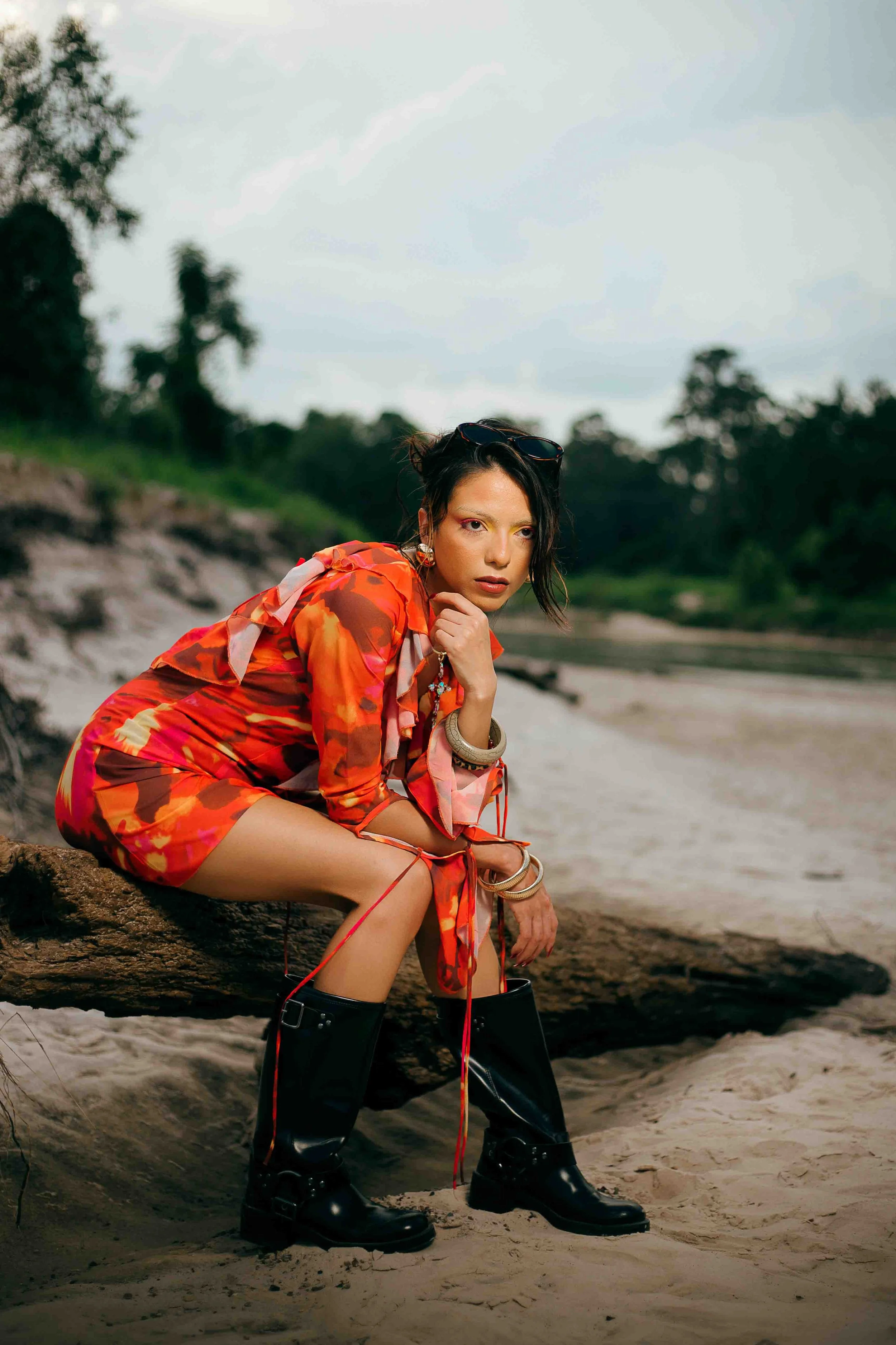Houston editorial portrait of a woman in a red patterned dress and black boots sitting on a fallen log by a sandy riverbank in a natural outdoor setting