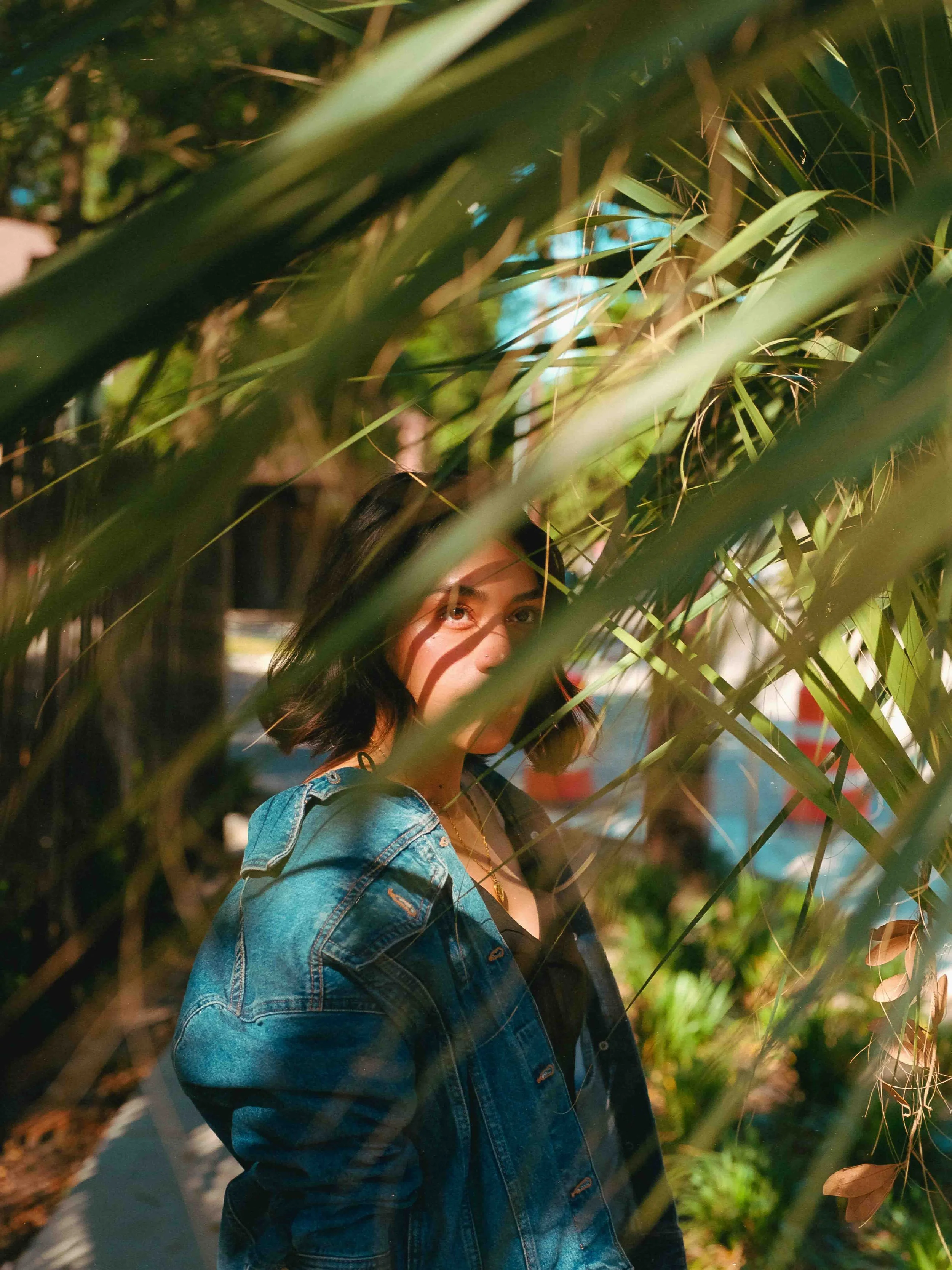 Film portrait of a young woman with short black hair wearing a denim jacket and black top, peeking through green foliage outdoors with natural light and soft 120 film tones kodak gold 200