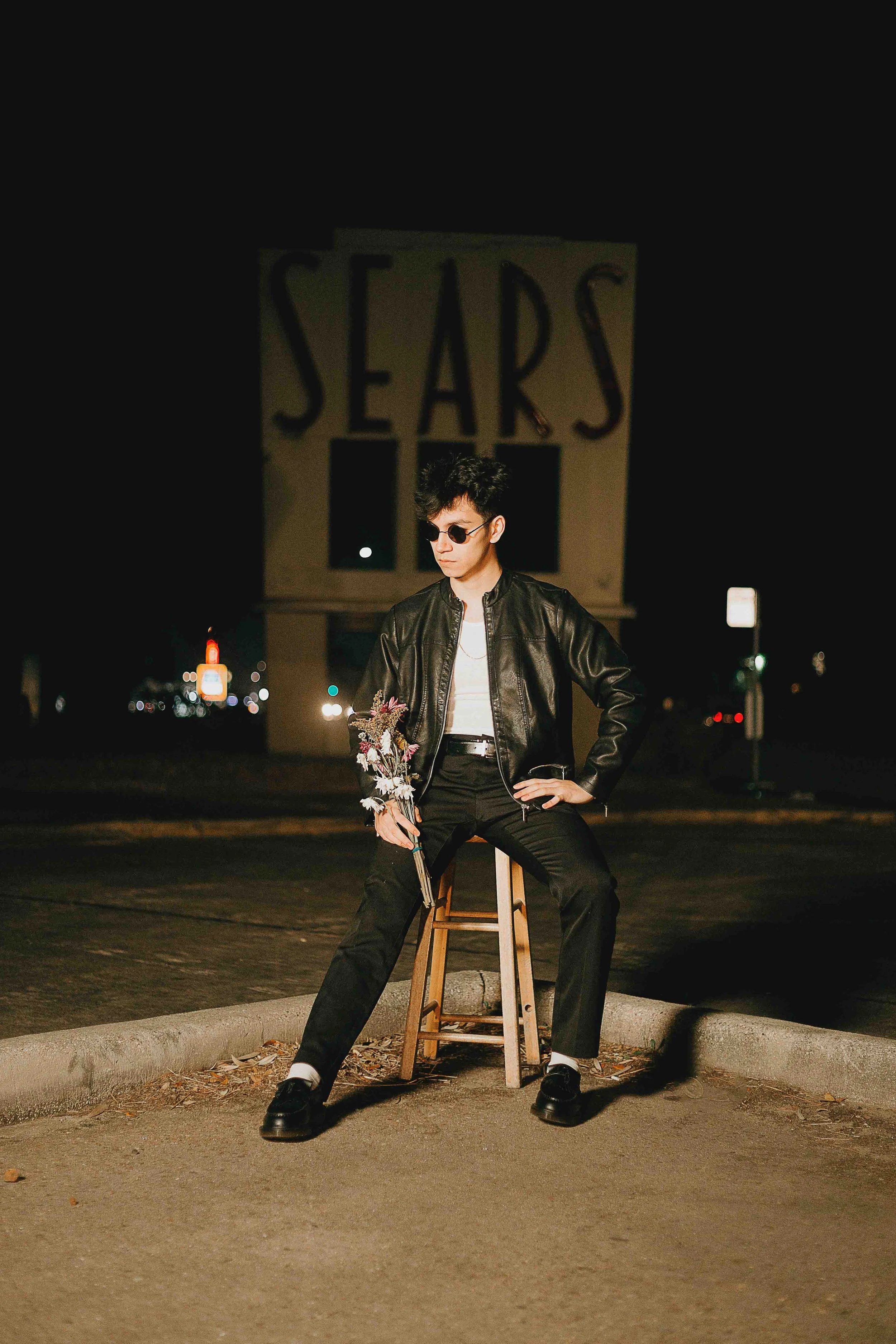 Houston editorial portrait of a person wearing a black leather jacket, sunglasses, and black pants sitting on a stool at night in front of a large ‘SEARS’ sign, holding a bouquet of flowers.