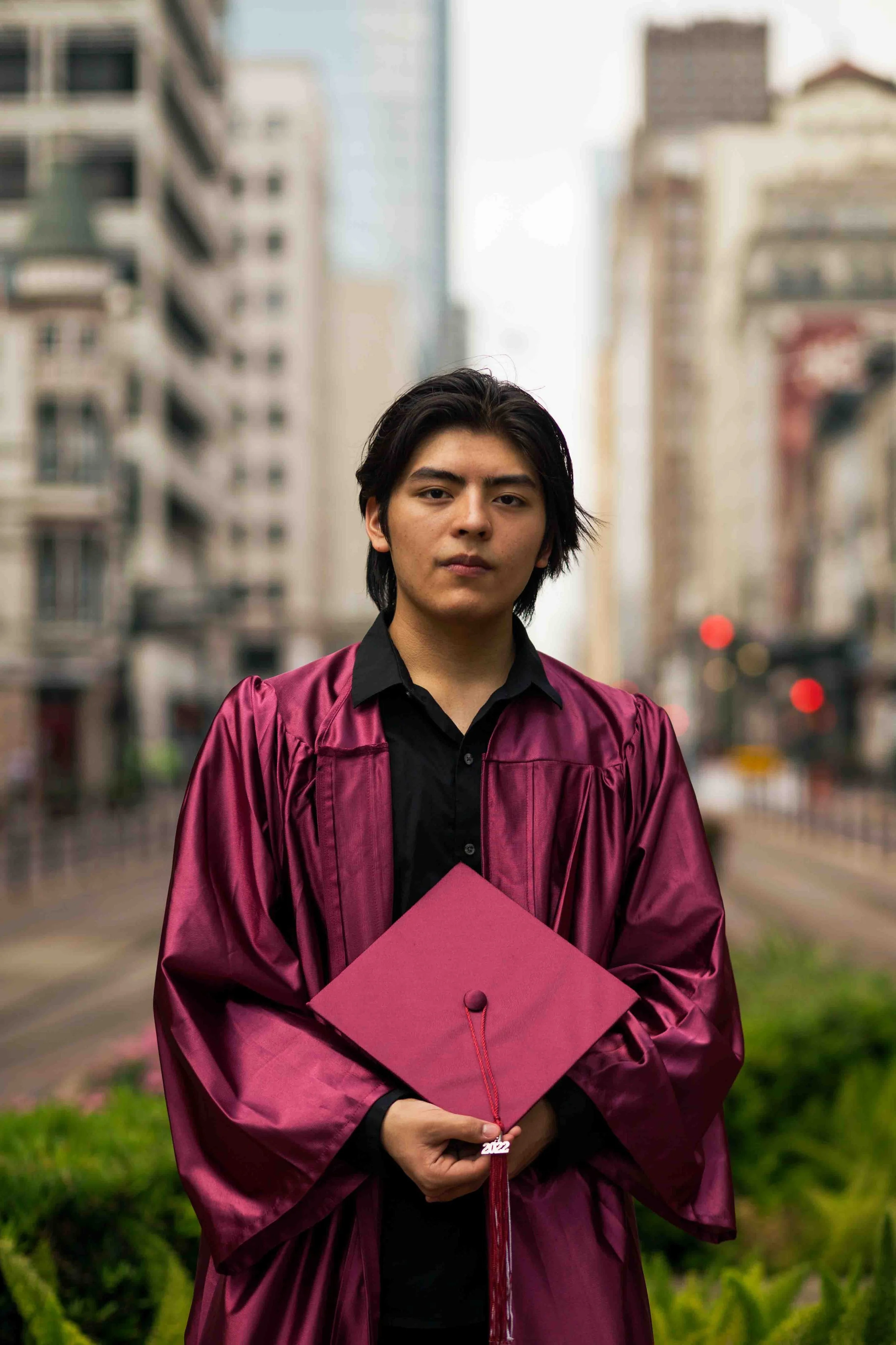 High school graduate wearing a maroon cap and gown, holding a mortarboard with a red “2022” tassel, standing in an urban setting with tall city buildings in the background