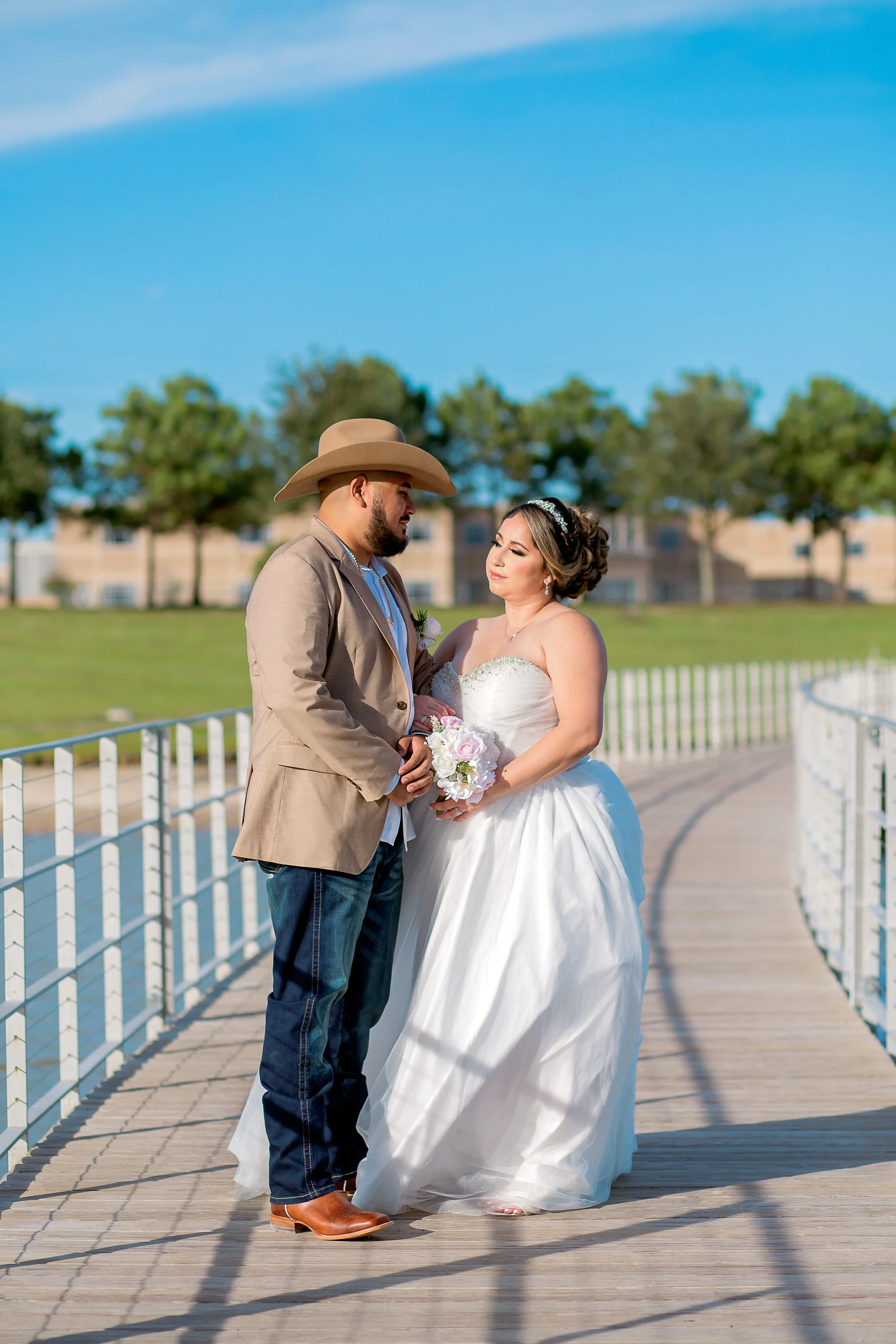 A bride and groom stand on a wooden bridge outdoors, holding hands, with the bride holding a bouquet of white and pink flowers, under a clear blue sky with trees in the background.
