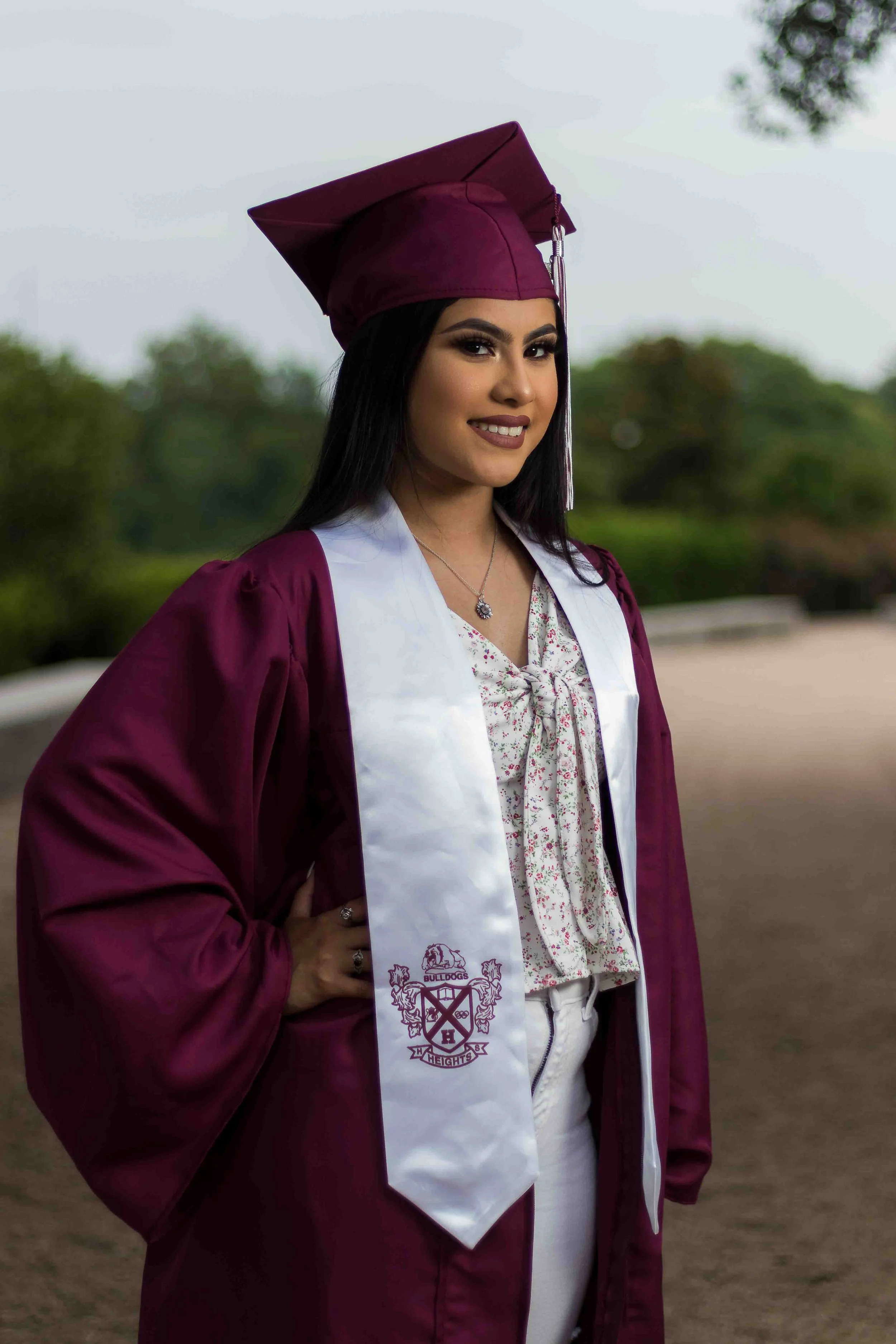 Houston Heights High School Class of 2020 senior graduation portrait by Lev's Photography featuring a young woman in a maroon cap and gown with a white sash and school crest, smiling outdoors with trees in the background in Houston, Texas.