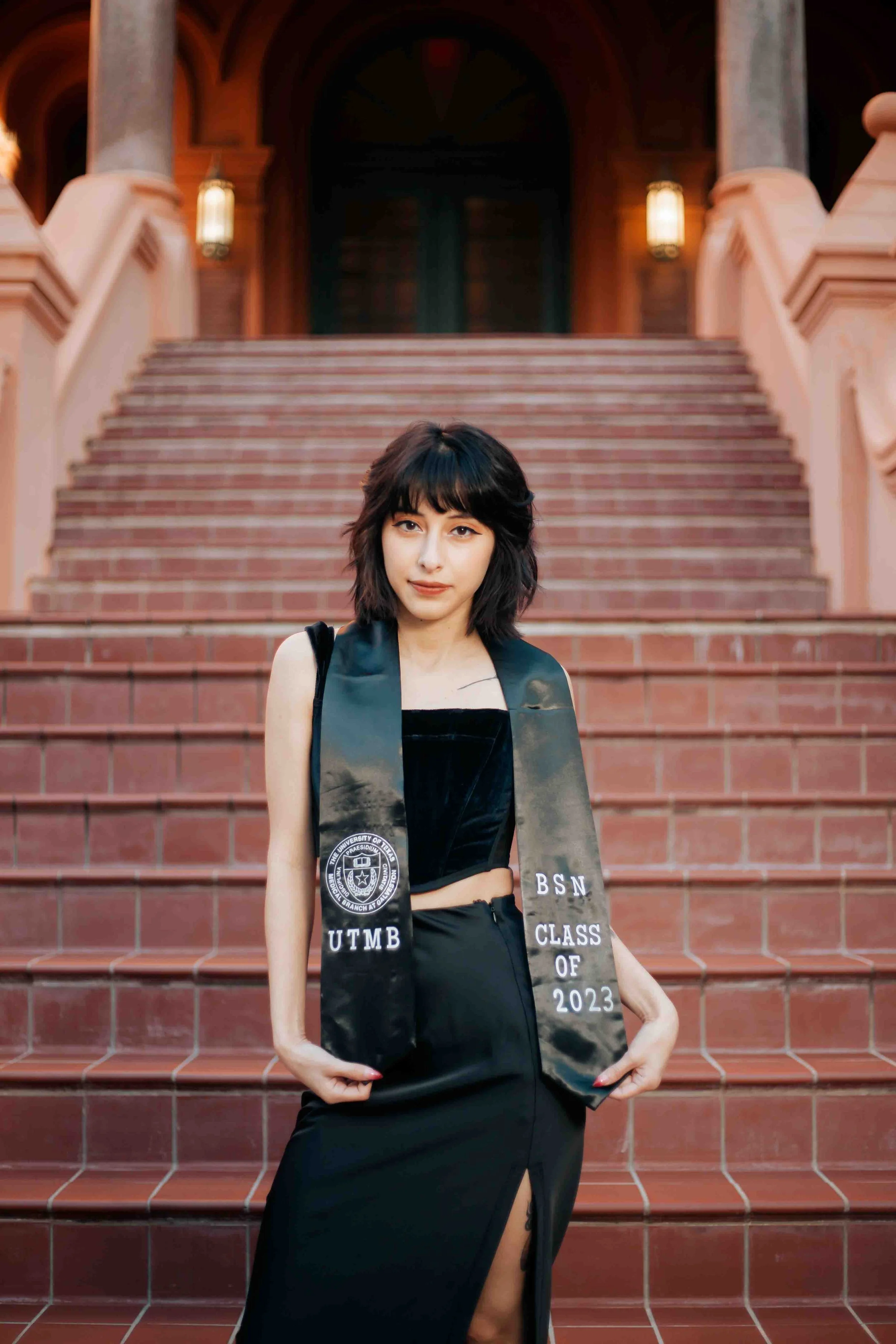 Graduation portrait of a young woman standing on red brick stairs outside a historic building, wearing a black dress and a graduation stole with university logos, celebrating her Class of 2023 achievement