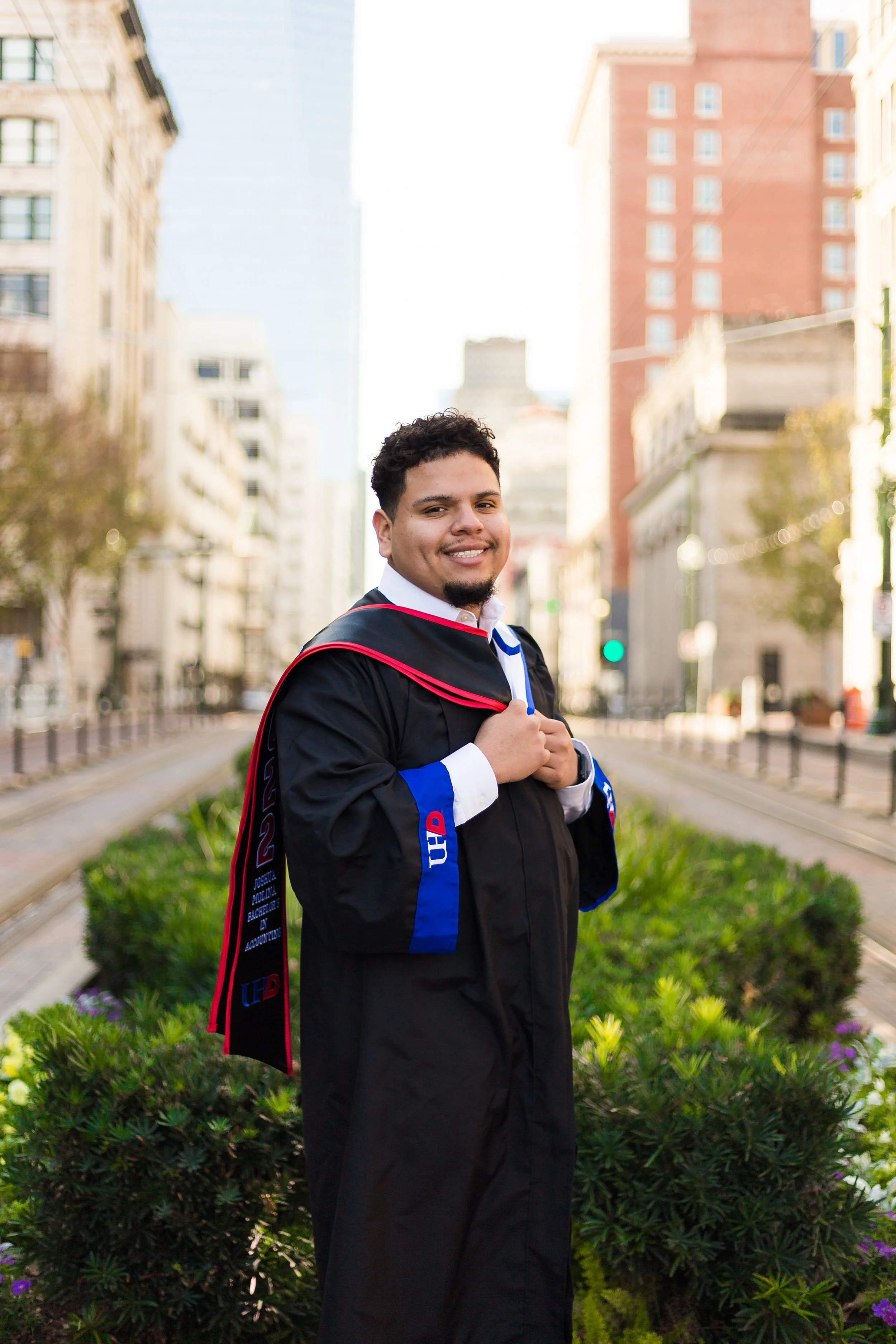A graduate in a black gown and academic hood standing outdoors on a city street, smiling with buildings and greenery in the background.