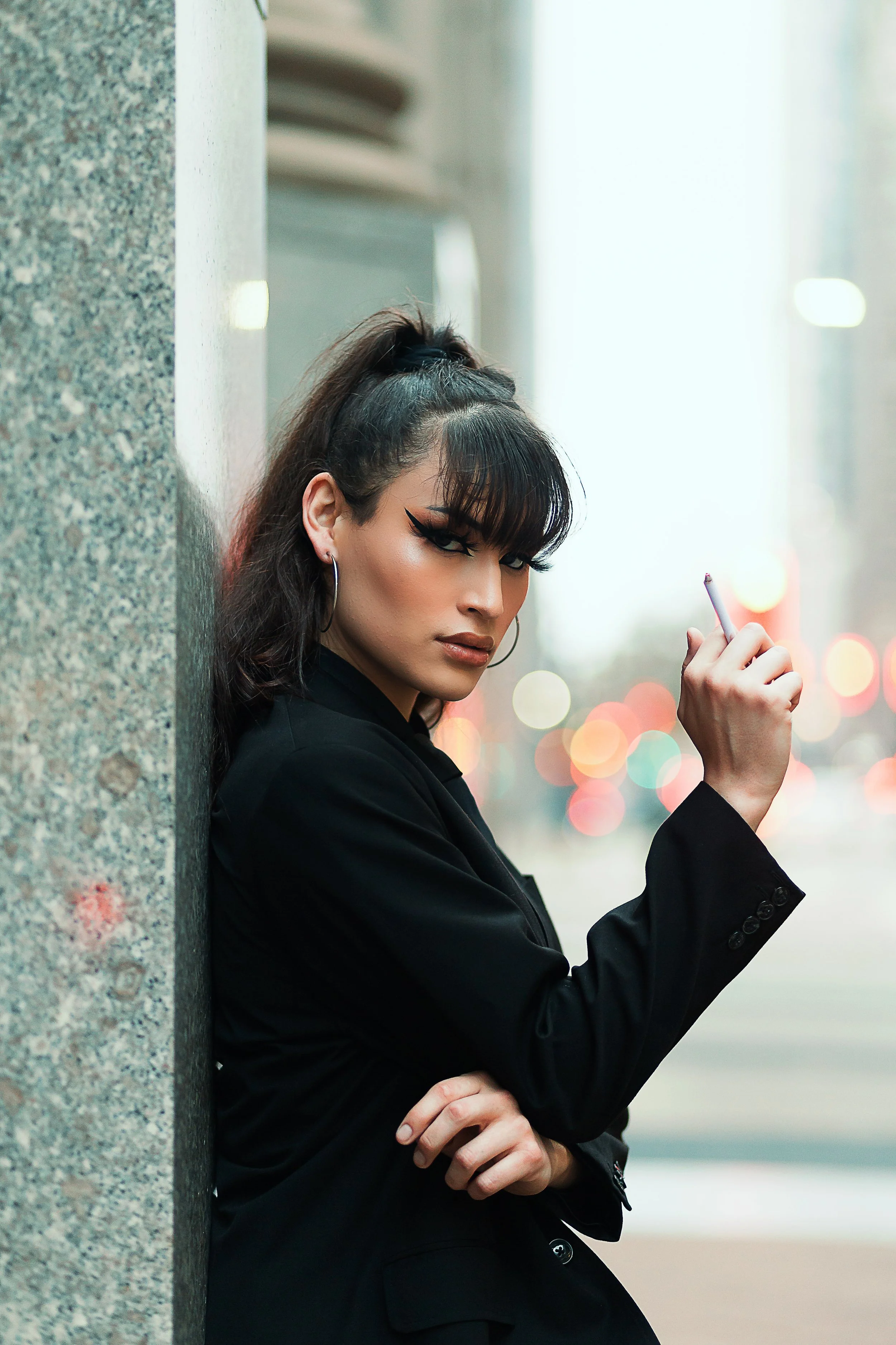 A woman with dark hair and makeup leaning against a concrete pole on a city street, holding a cigarette in her right hand, dressed in a black blazer.