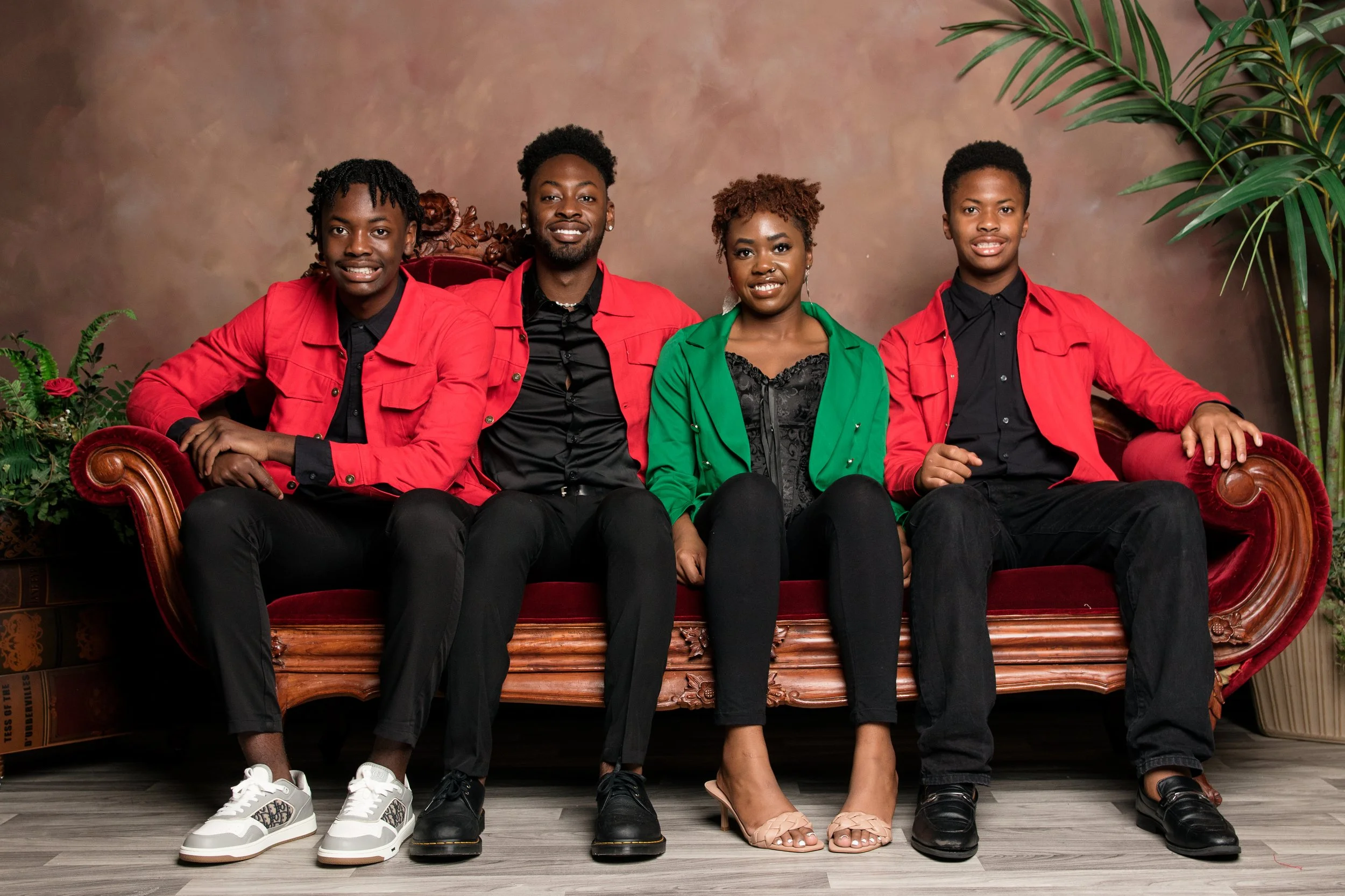 Four young adults sitting on a vintage red velvet sofa in front of a brown backdrop, with green plants on either side. They are dressed in black pants and black tops, with some wearing red or green jackets. They are smiling at the camera.
