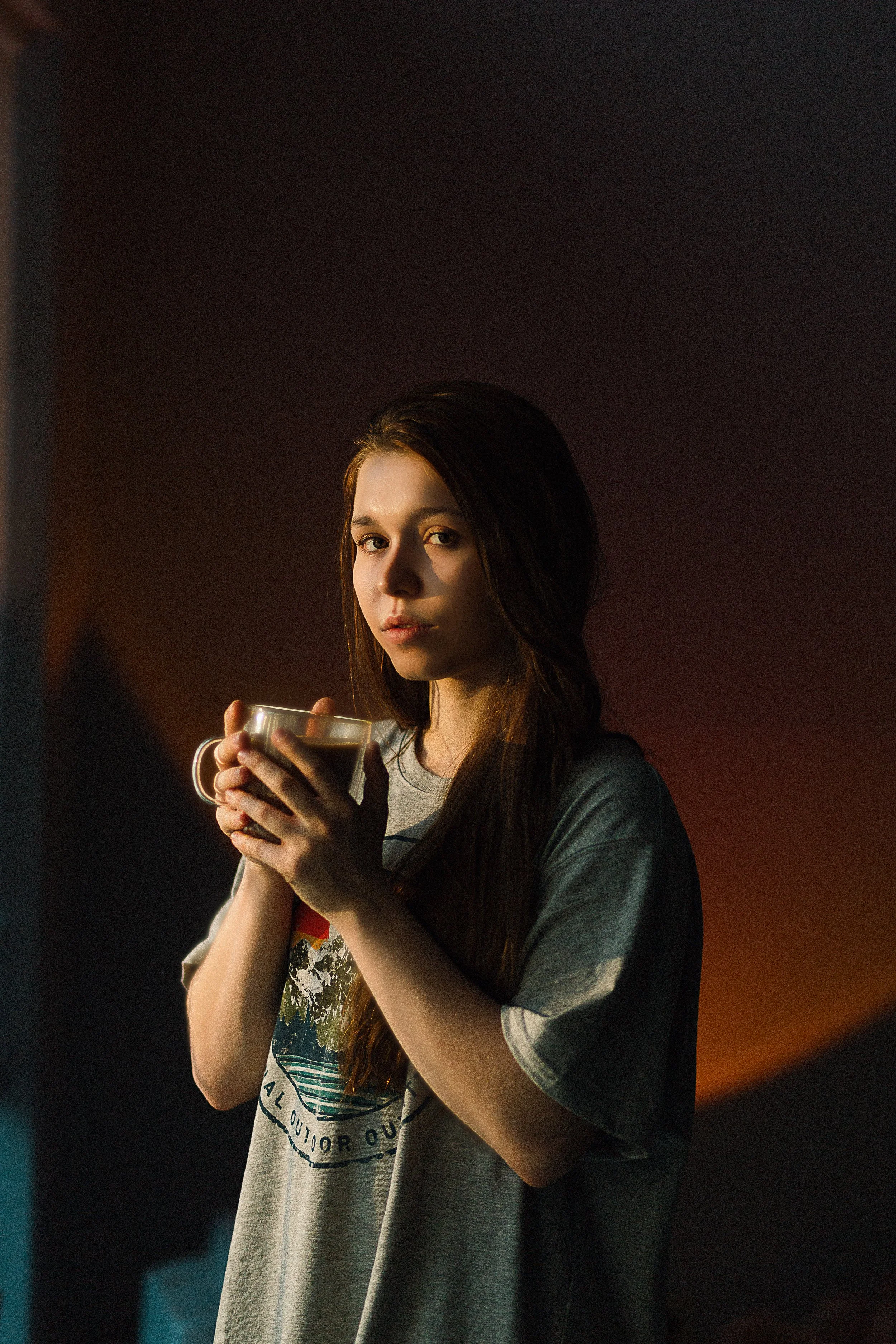 A young woman with long brown hair holding a coffee mug, looking at the camera while standing indoors with dim lighting.