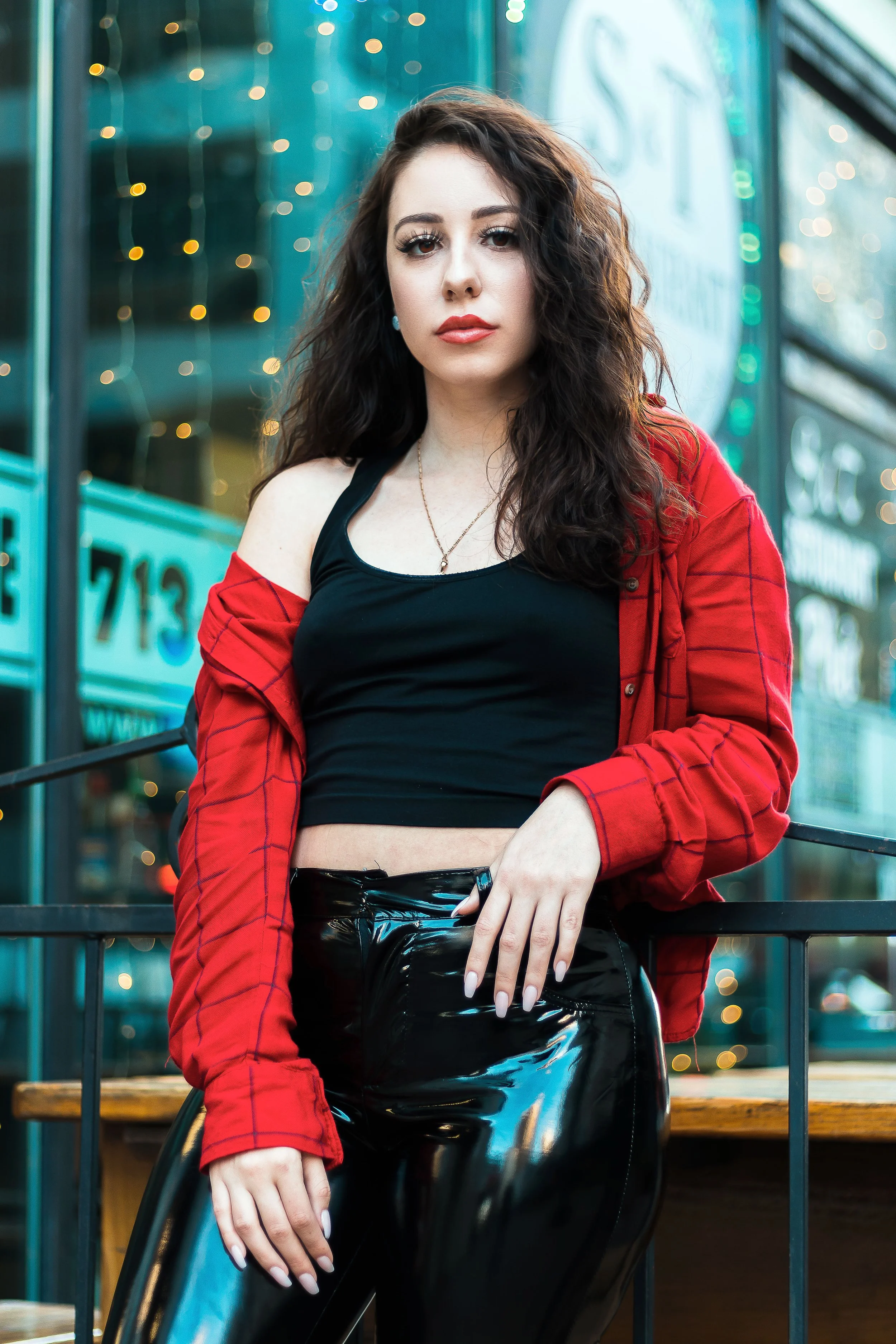 A young woman with dark, wavy hair and makeup, wearing a black crop top, shiny black pants, and a red jacket, standing outdoors near a railing in a city setting with blurred lights in the background.