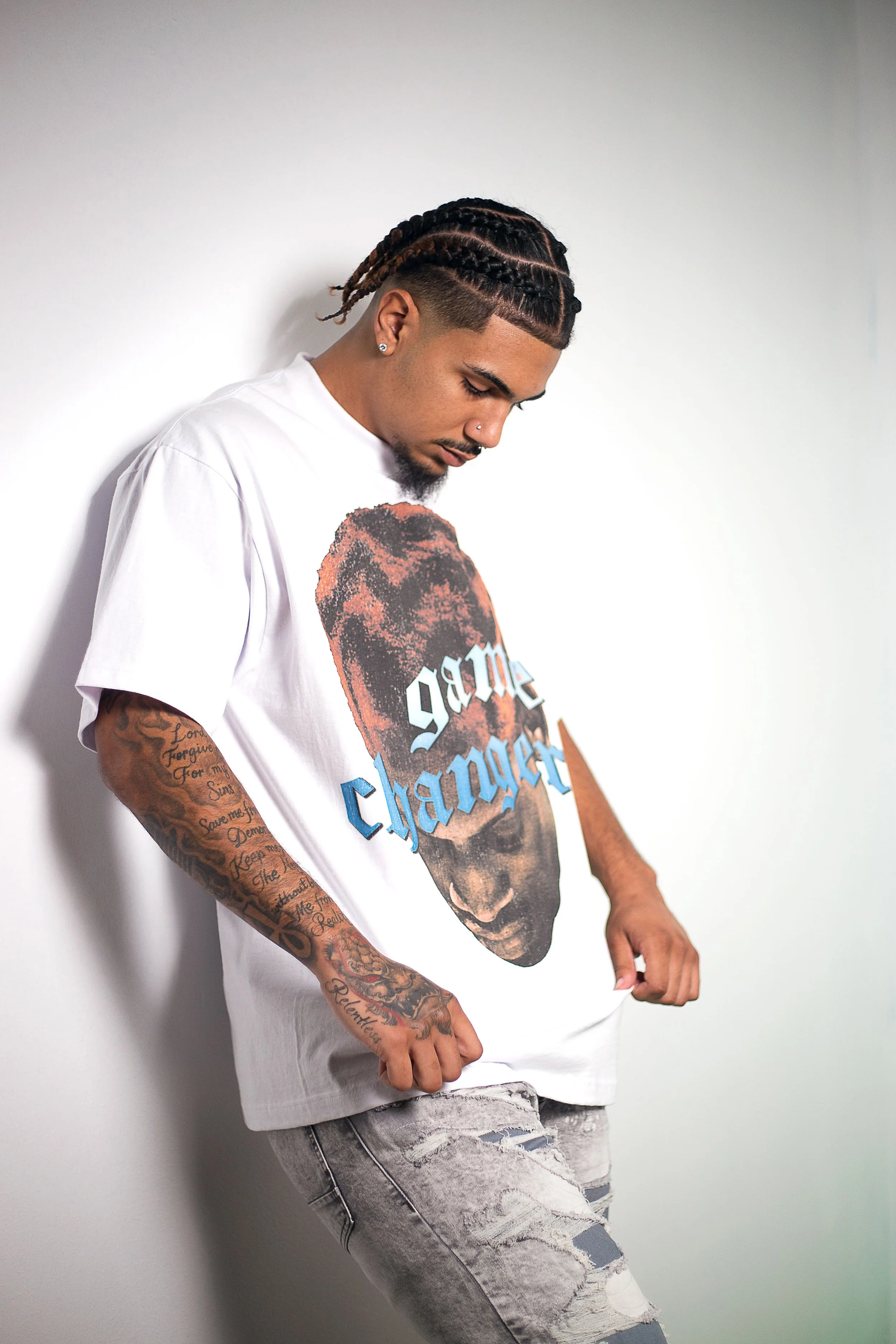 Young man with braided hair and tattoos, wearing a white T-shirt with a graphic print of a woman's face and text, and ripped jeans, standing against a plain white wall.