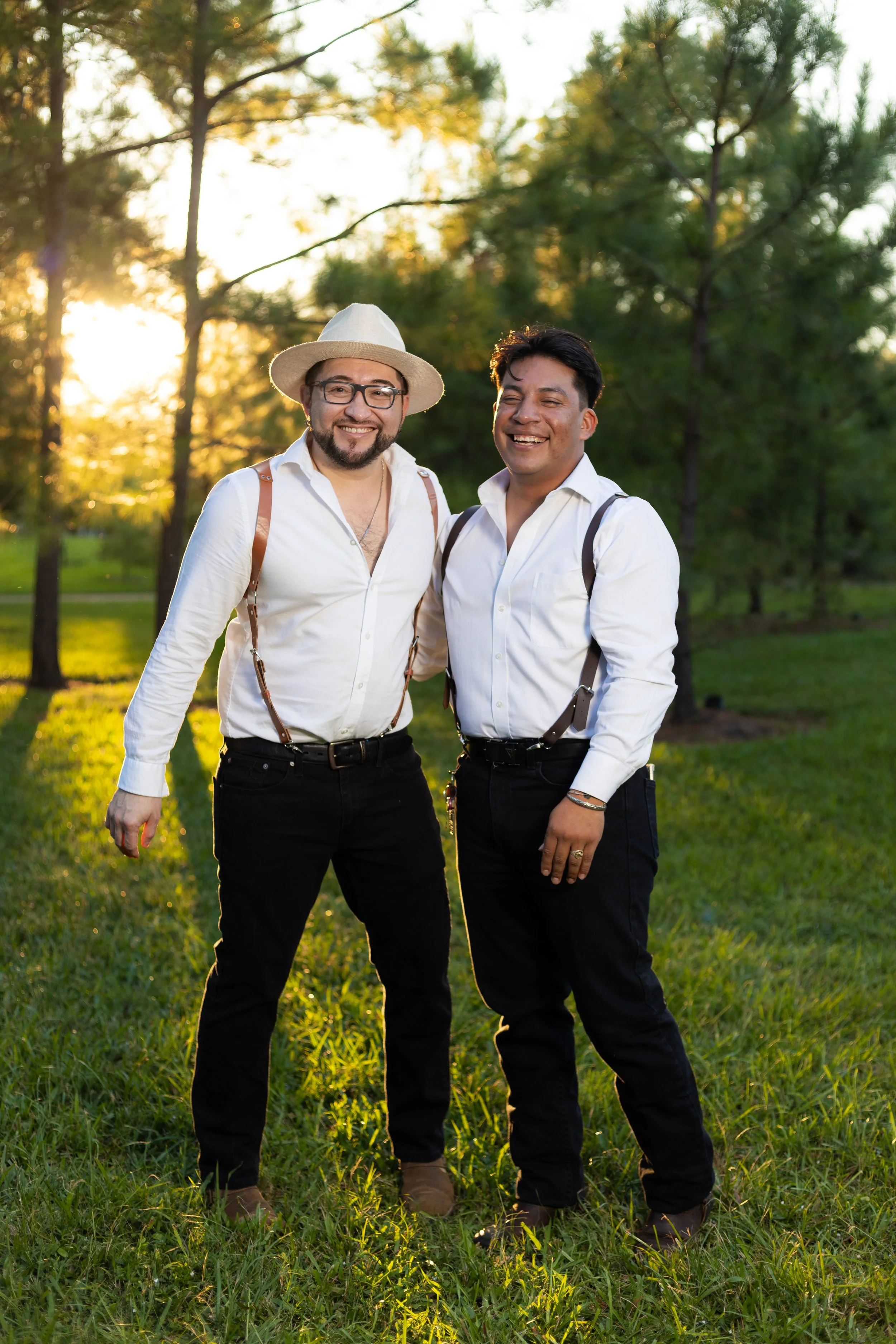 Two men standing together outdoors in a park during sunset, smiling, wearing white shirts, black pants, suspenders, and brown shoes.