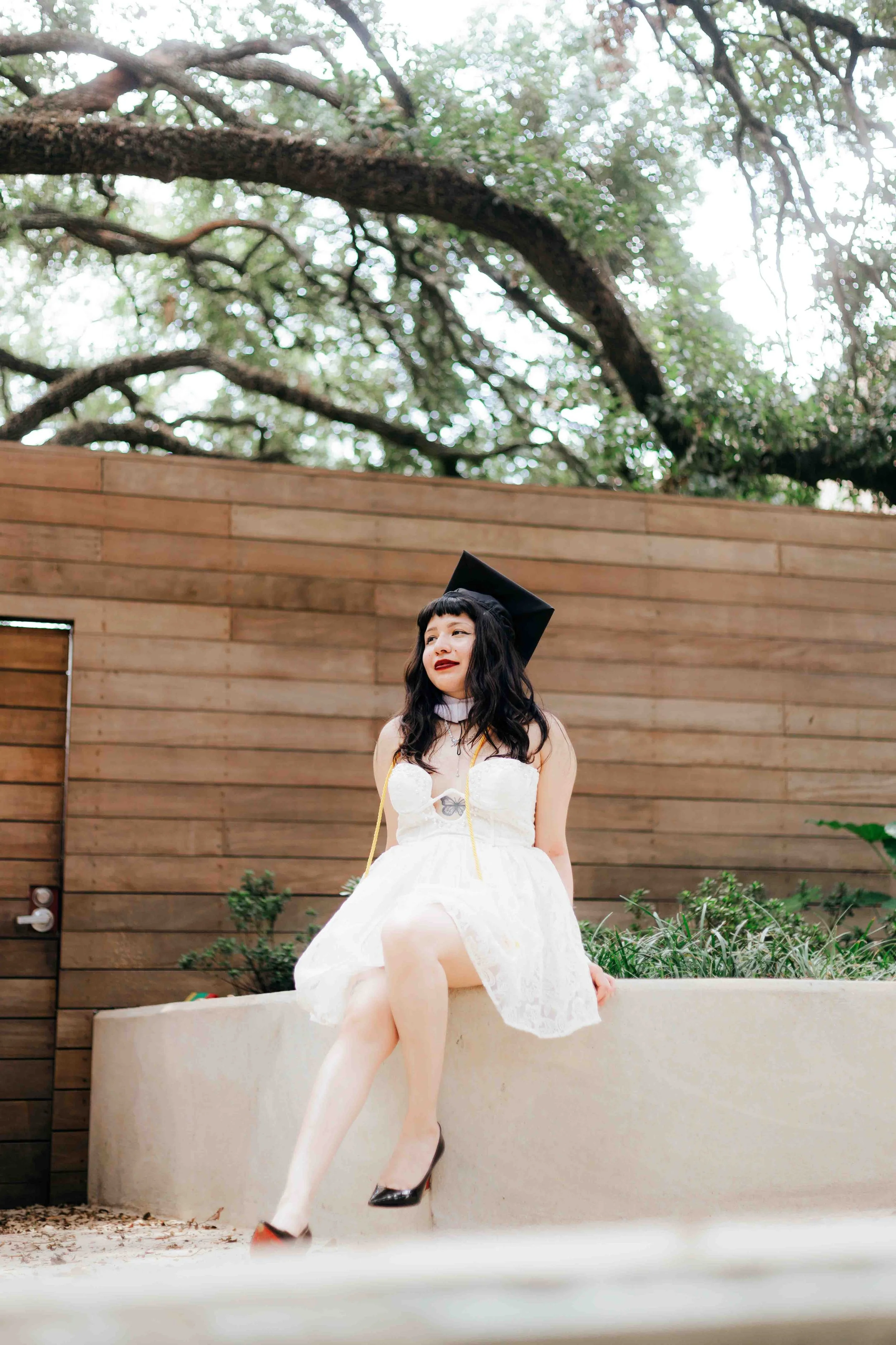 Graduation portrait at Rice University in Houston featuring a smiling young woman in a white dress and black cap sitting on a low wall with trees and a wooden fence in the background