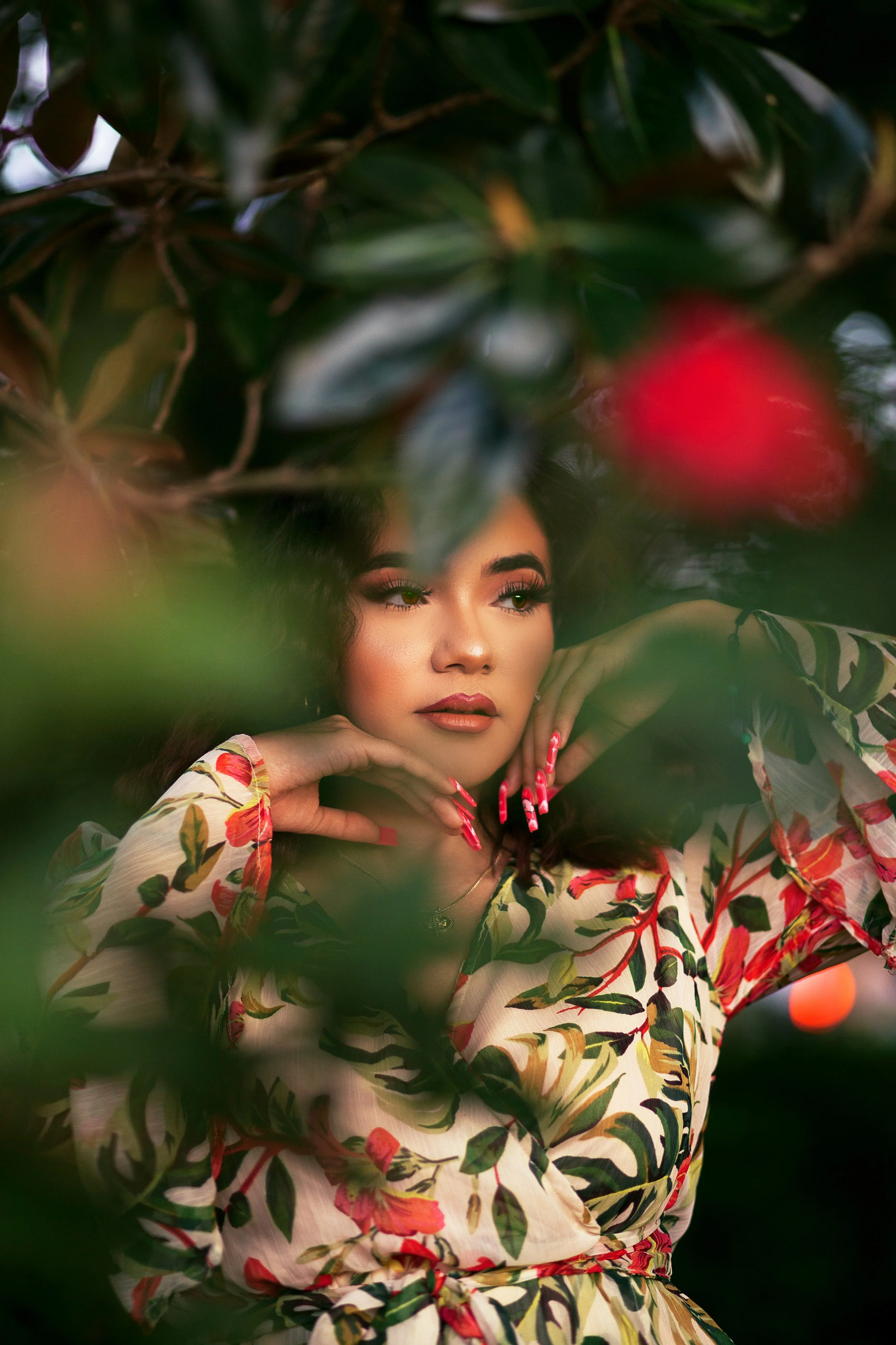 A woman with long dark hair and makeup posing among green leaves and foliage, wearing a colorful floral top with red, green, and white patterns.