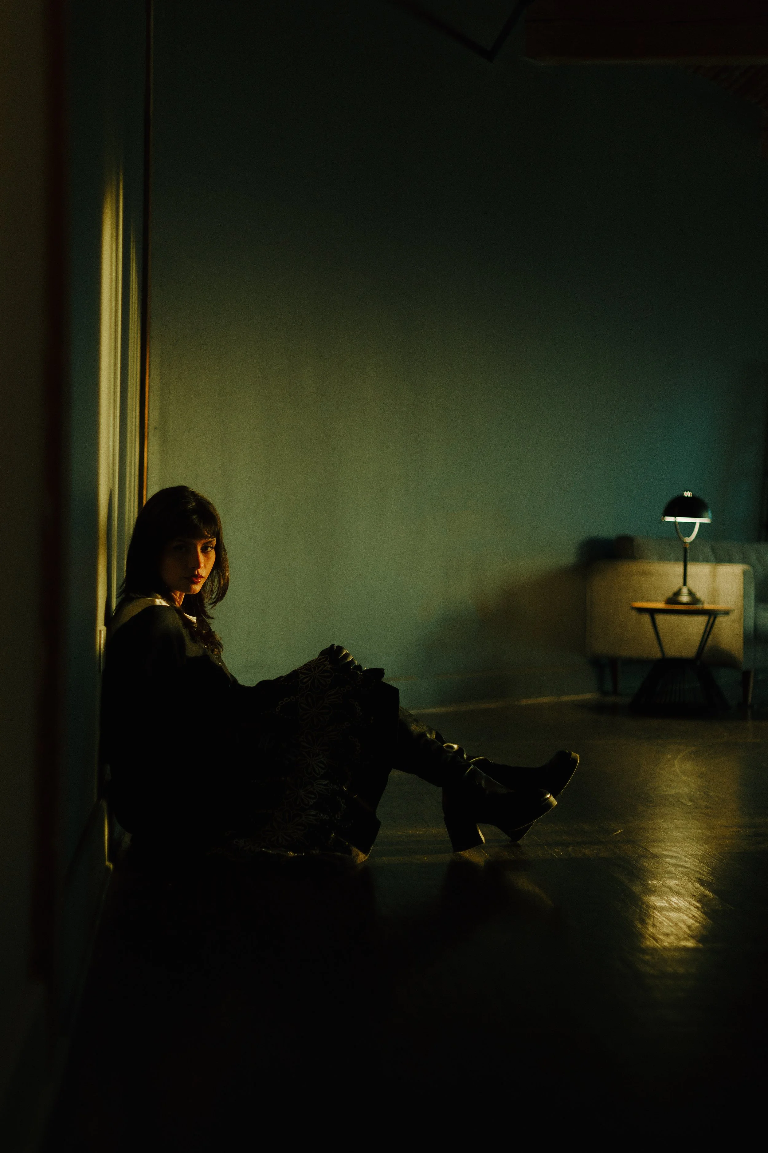 A woman with dark hair sitting on the floor in a dimly lit room, illuminated by a soft light from the left, with a sofa and table with a lamp in the background.
