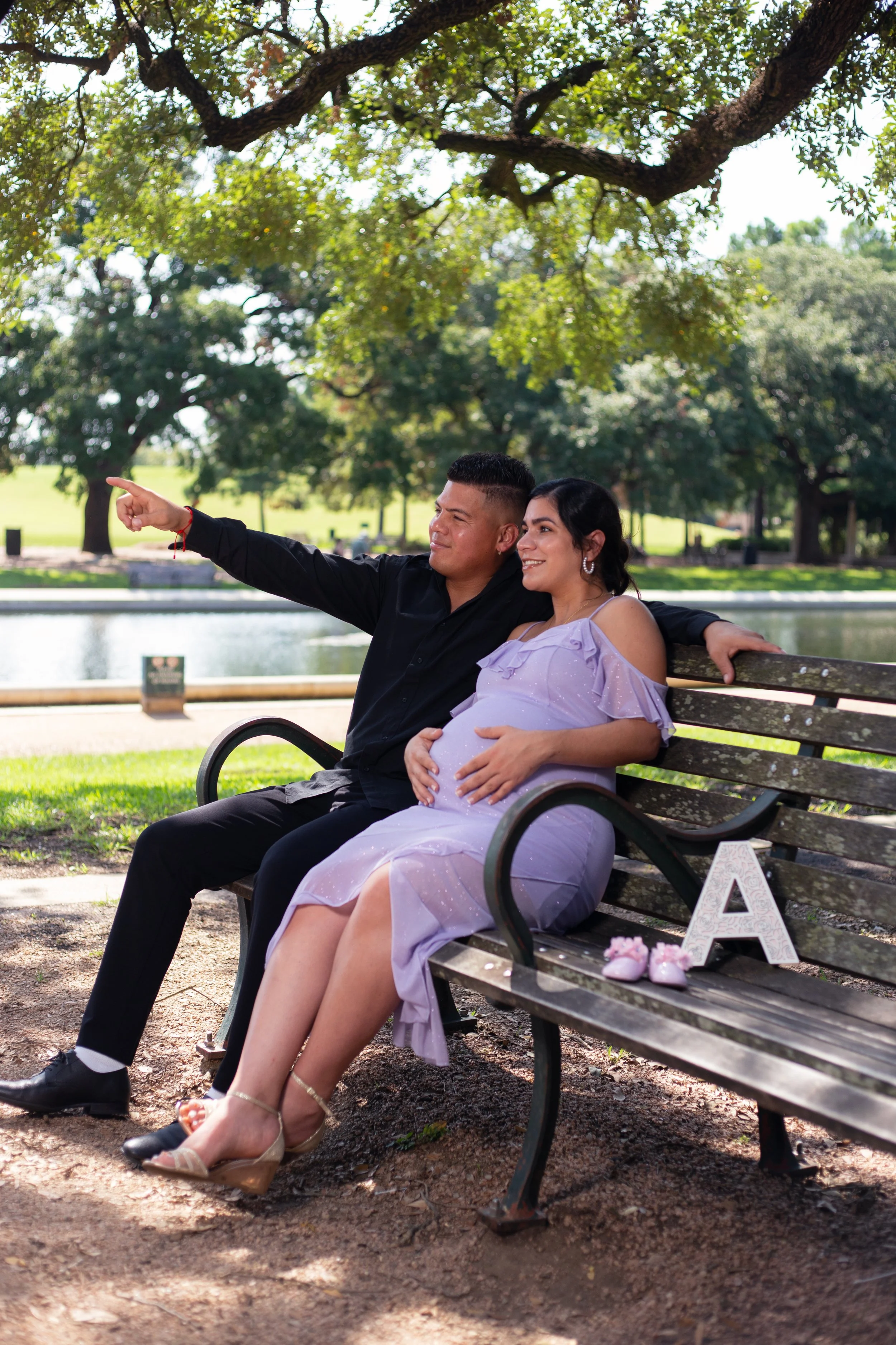 Couple sitting on park bench, woman pregnant, man pointing, lakeside background with trees, pink baby shoes and letter 'A' on the bench.