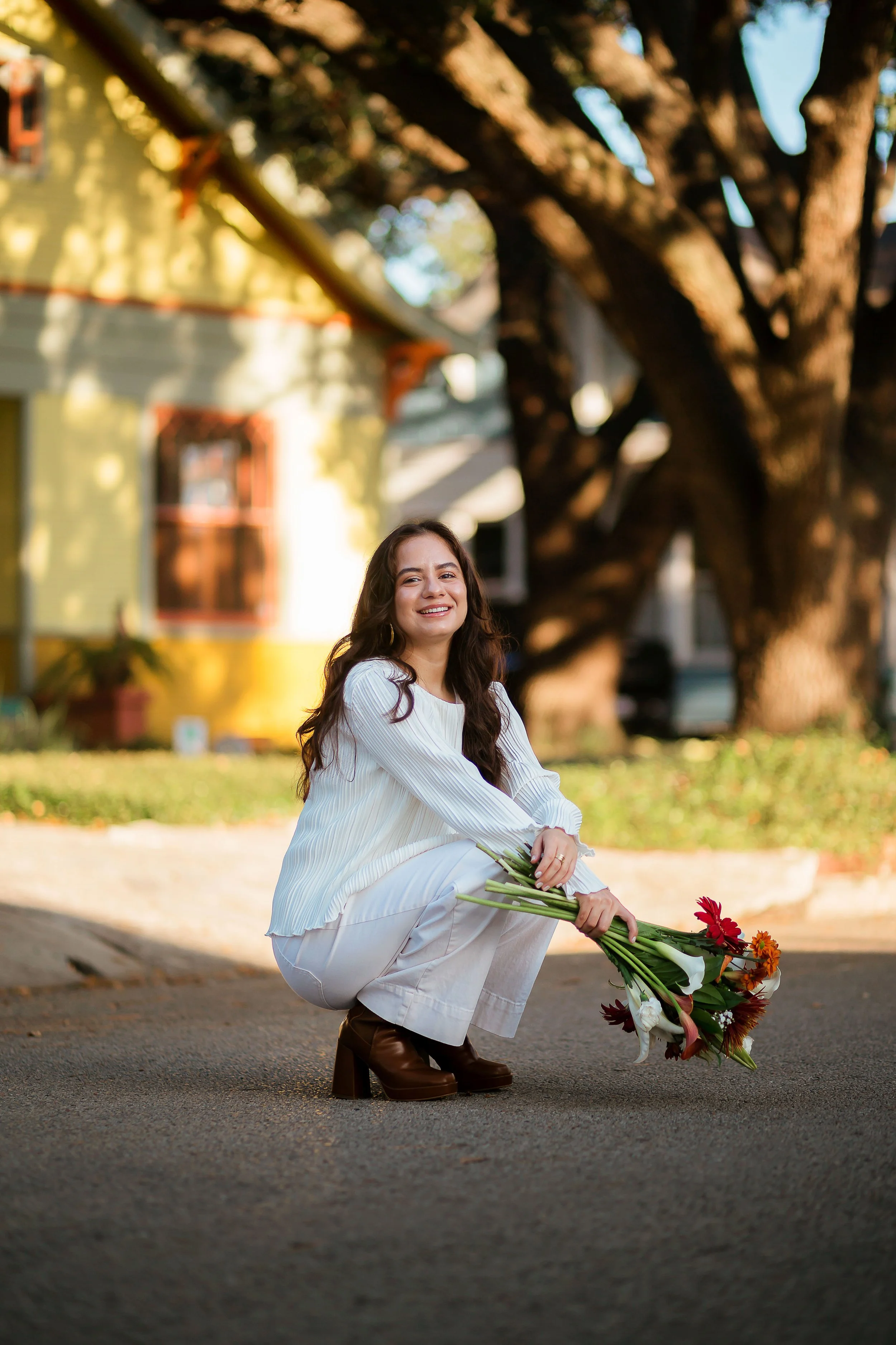 A woman with long dark hair smiling and holding a bouquet of flowers while squatting on a street in front of a large tree and yellow house.