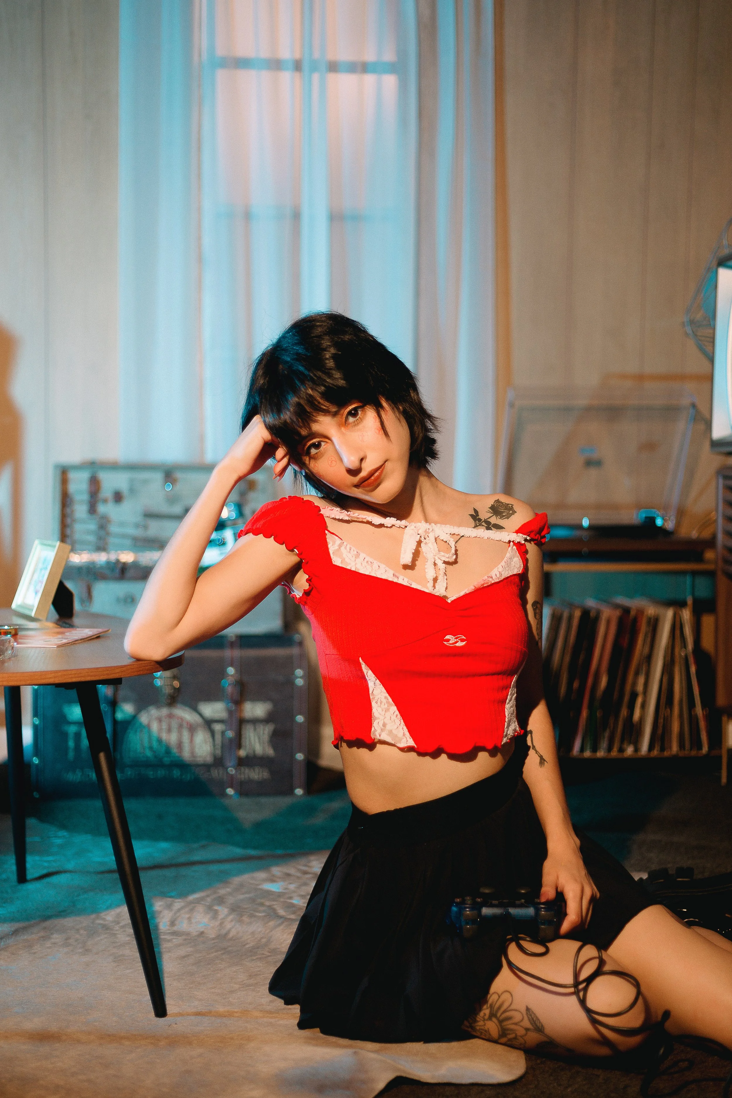 A young woman with black hair and tattoos, wearing a red crop top and black skirt, sitting on the floor in a room with vinyl records and a turntable in the background.