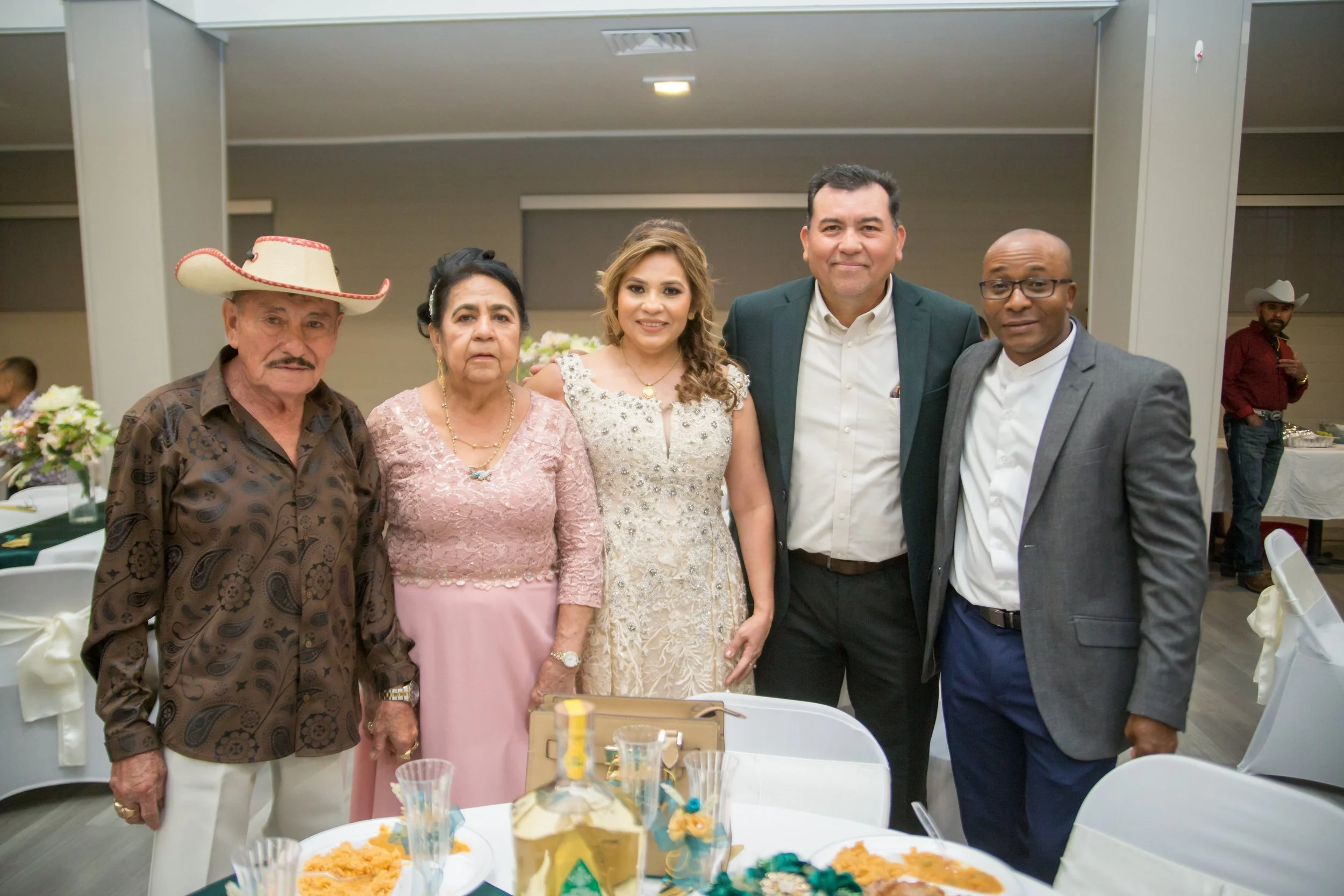Group of six people, including an elderly man in a cowboy hat, an elderly woman, a young woman in a formal dress, and three men in suits, standing together at a celebration or social event, with tables and food visible in the foreground.