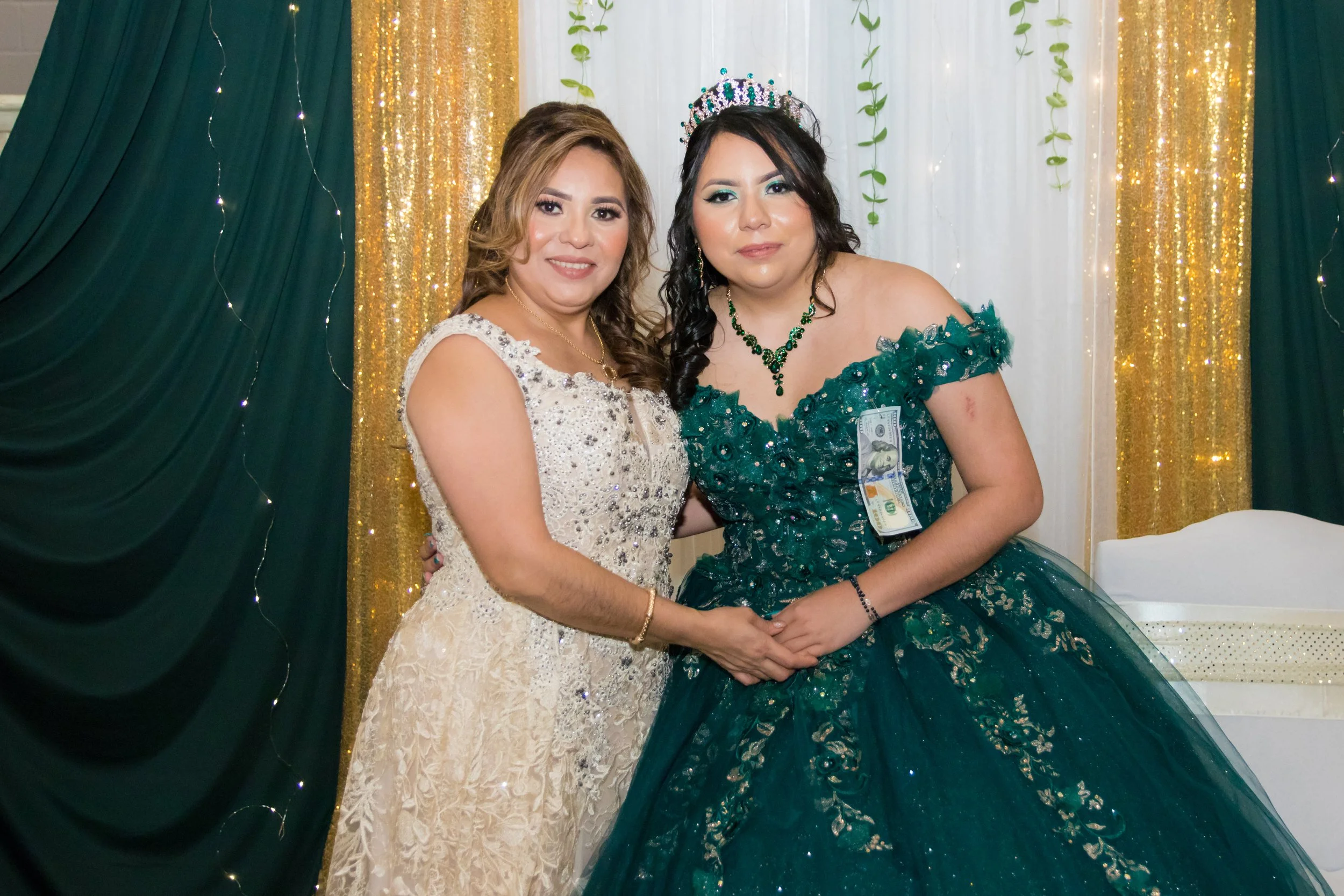 Two women in formal gowns standing together at a celebration, one wearing a white dress and the other in a green gown with a tiara, against a backdrop of green and gold curtains.