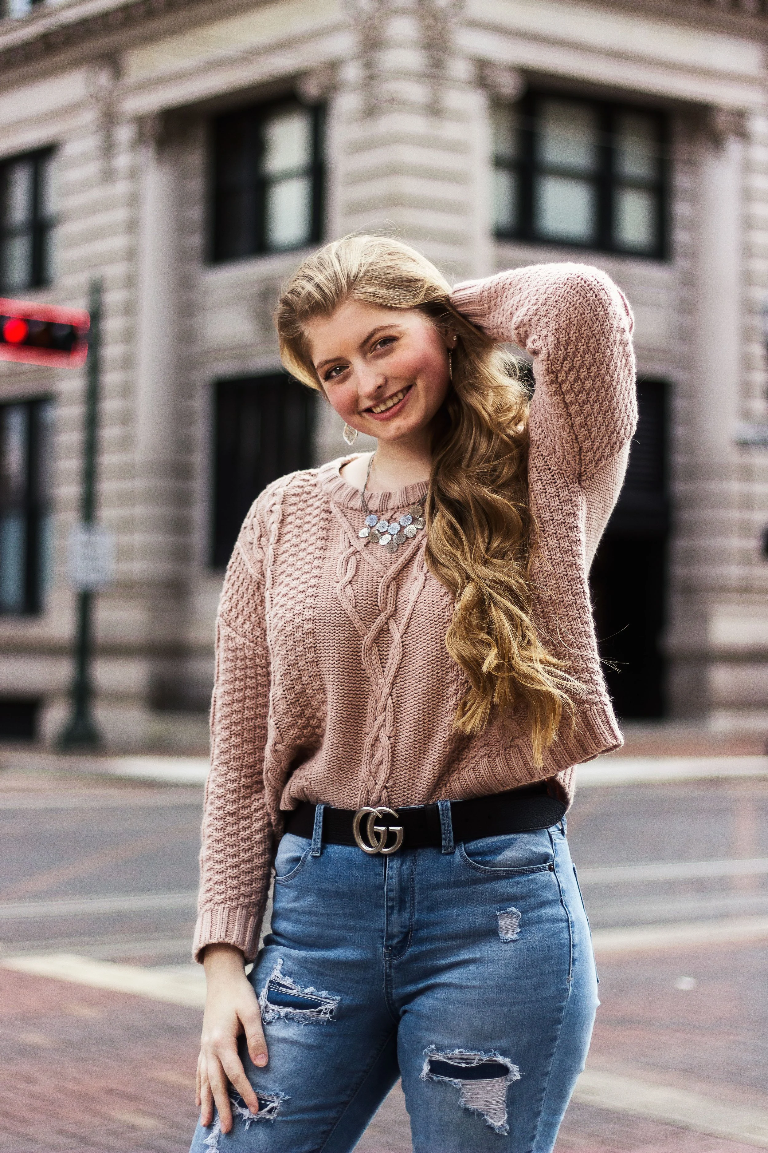 A young woman with long, wavy blonde hair smiling and posing outdoors in front of a city building. She is wearing a pink knitted sweater, distressed jeans, and a black belt with a Gucci logo.