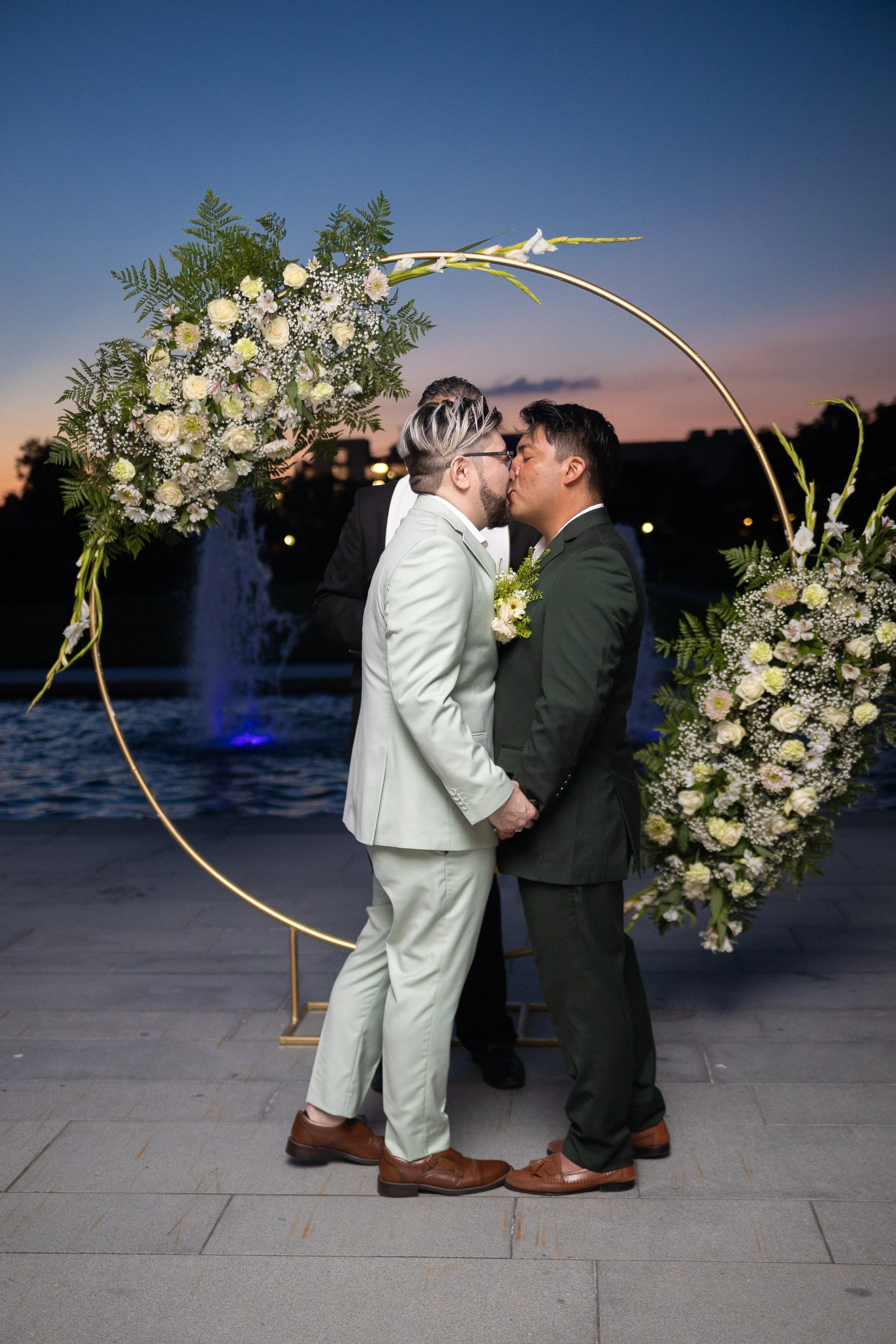 Two men in suits kiss during their wedding ceremony at sunset with a floral arch and a fountain in the background.