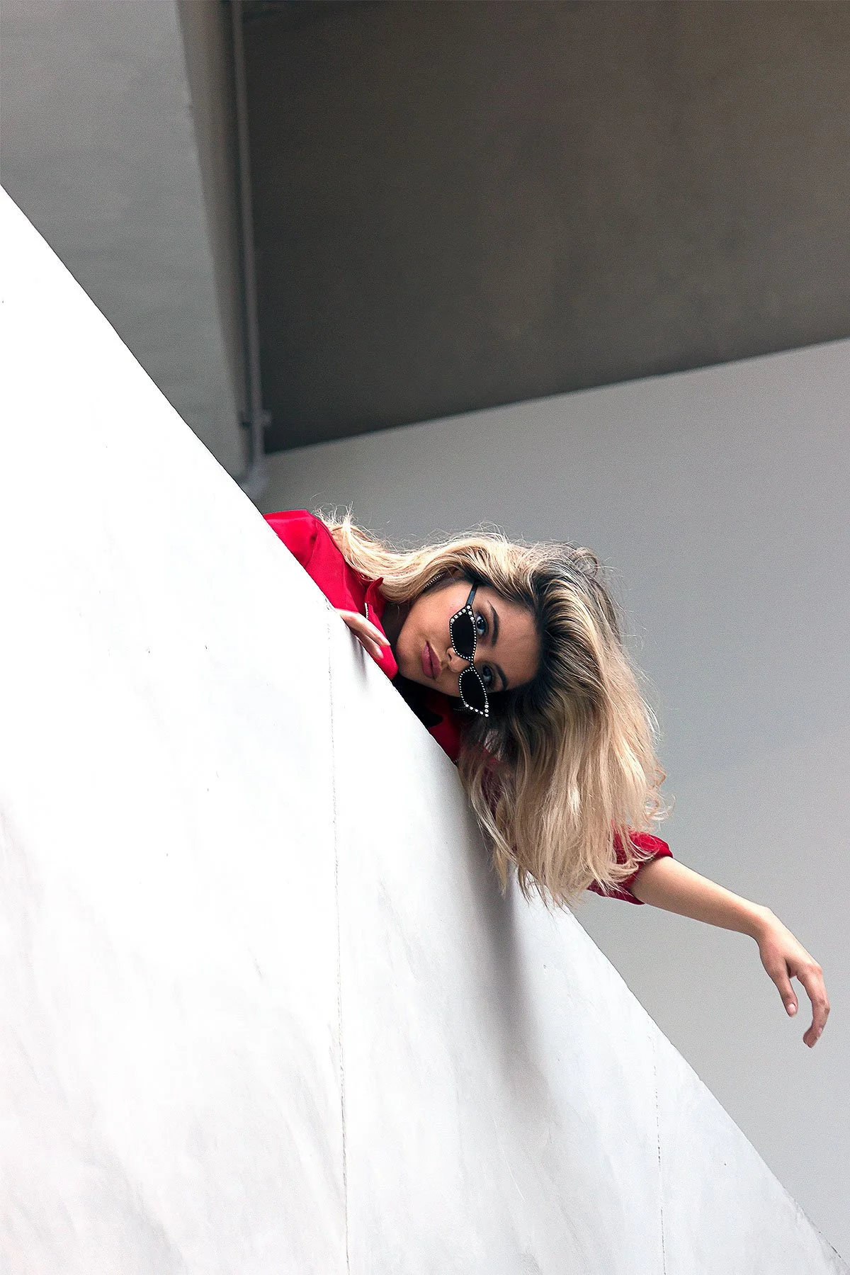 Editorial fashion portrait in Houston, Texas by Lev’s Photography featuring a blonde woman in sunglasses and a red jacket leaning over a white wall during a creative outdoor session
