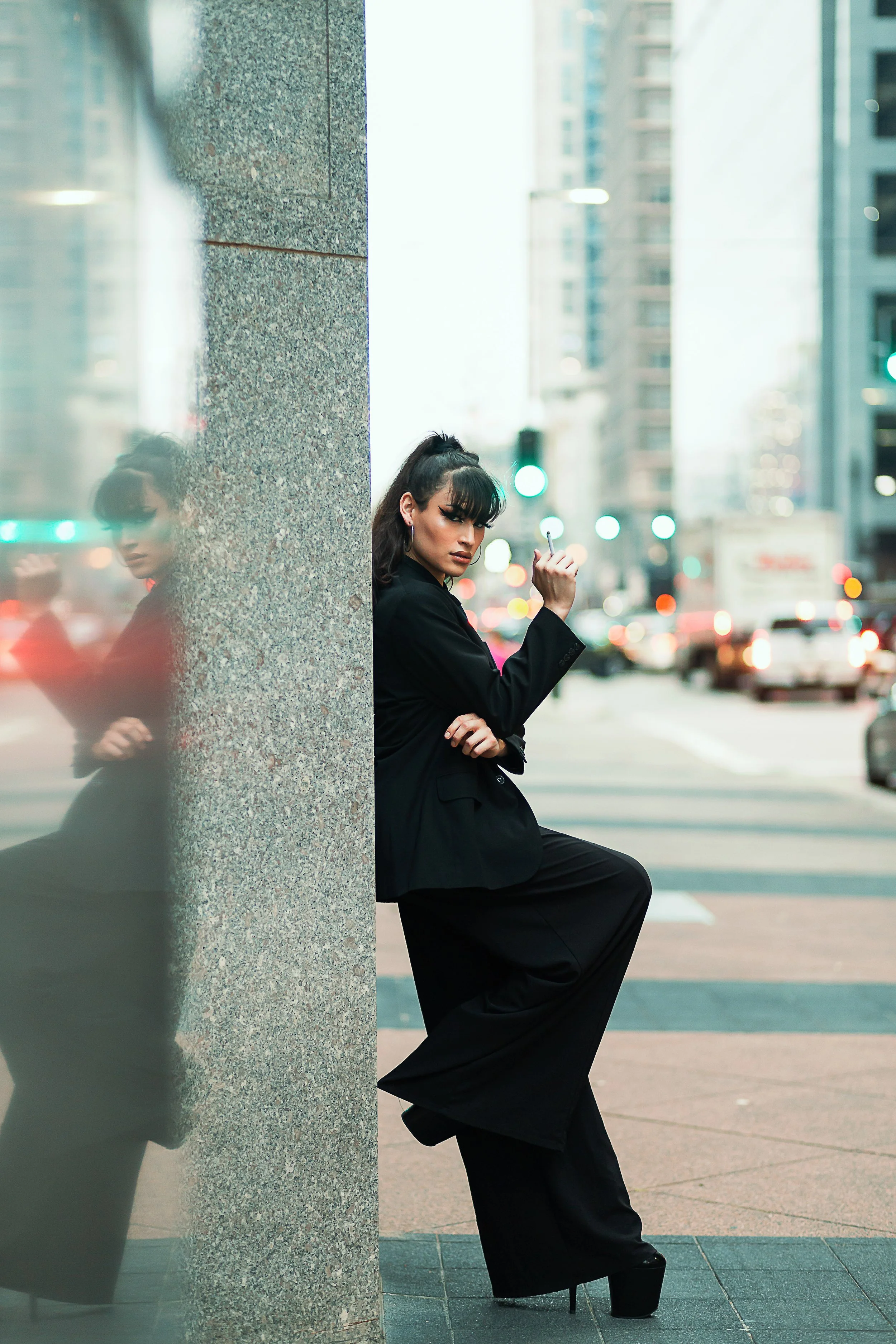 A woman in black business attire leaning against a building corner on an urban street, with her reflection visible in a glass surface nearby.