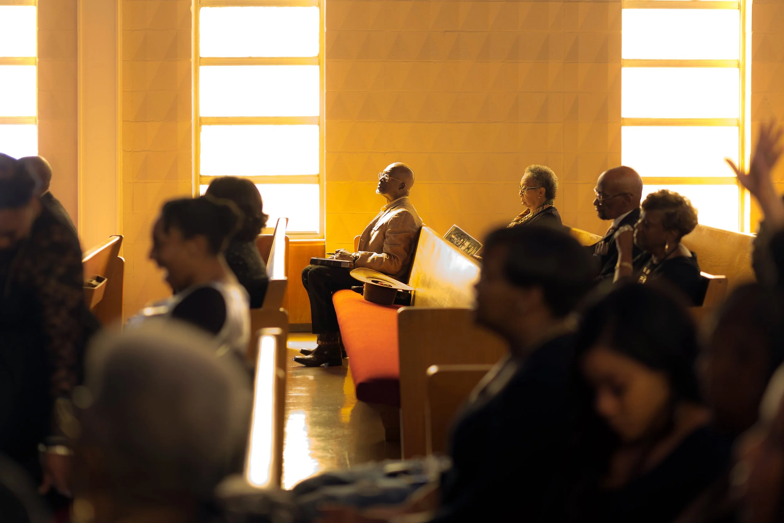 People sitting in pews during a church service or event, with sunlight streaming through large windows in the background.