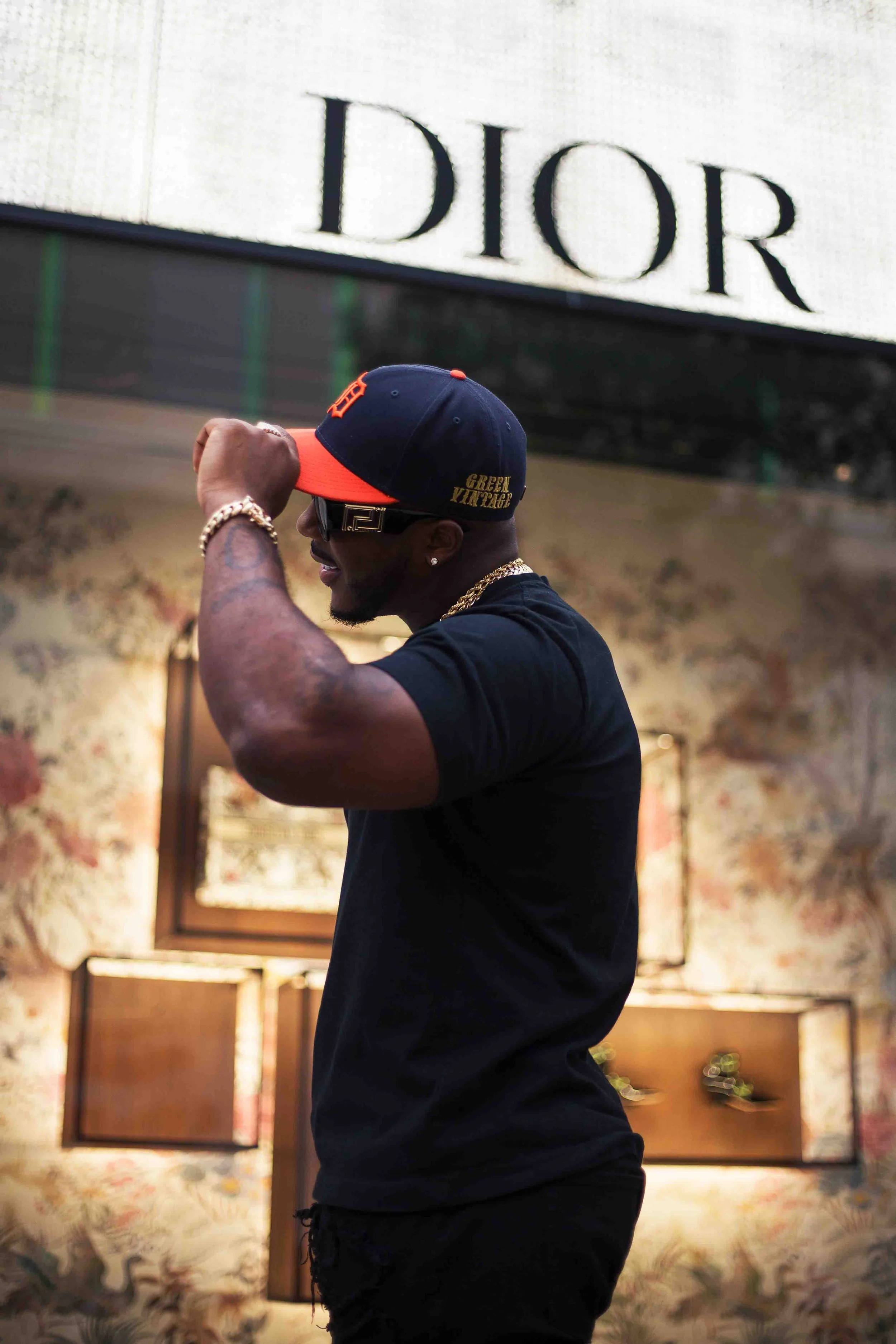 Luxury retail branding portrait in Houston, Texas by Lev’s Photography featuring a man wearing a dark baseball cap, sunglasses, gold chain, and black T-shirt adjusting his cap inside a store with floral wallpaper, shoes on display, and a visible Dior