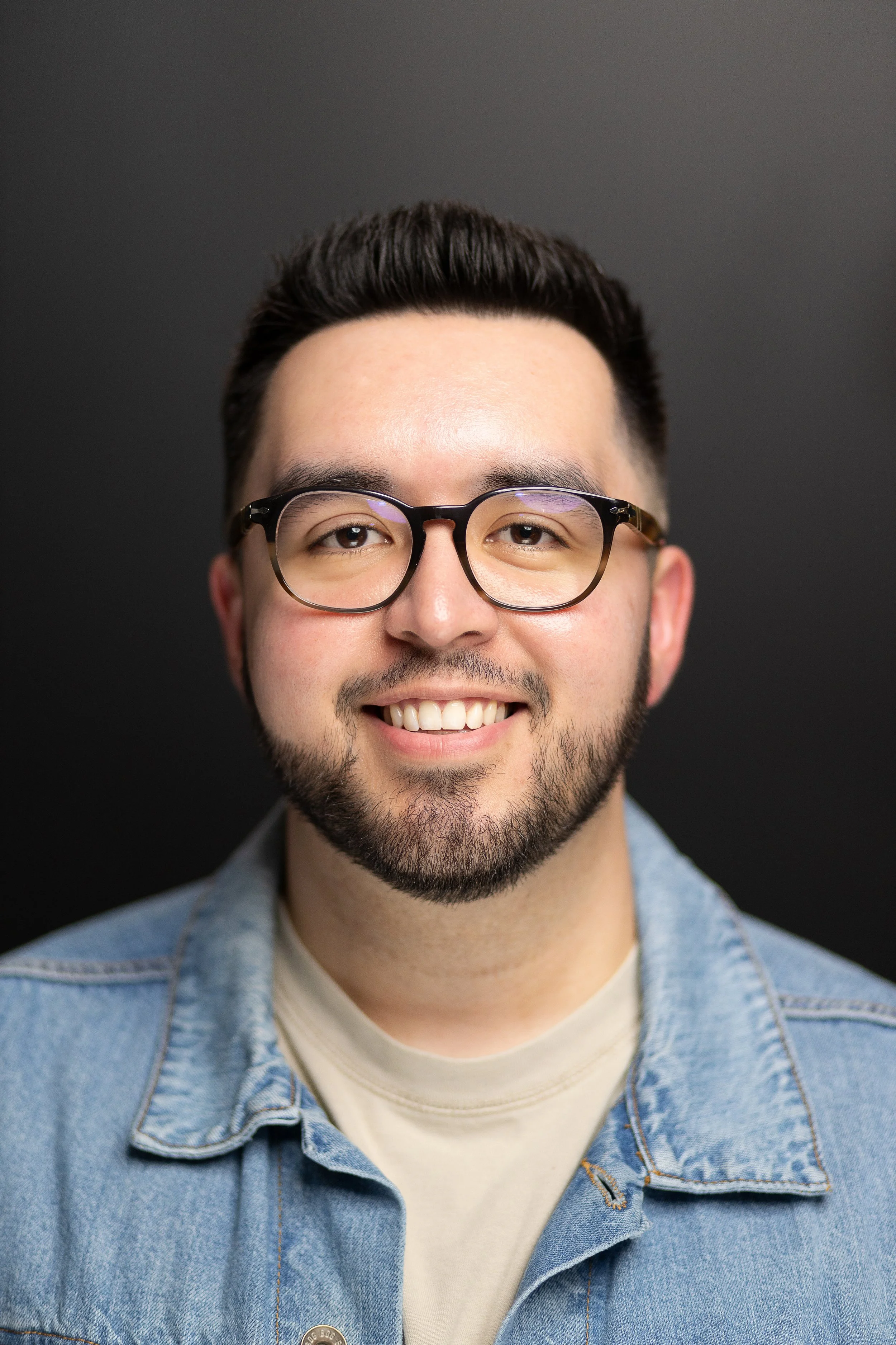 Close-up portrait of a young man with glasses, smiling, wearing a denim jacket, against a dark background.