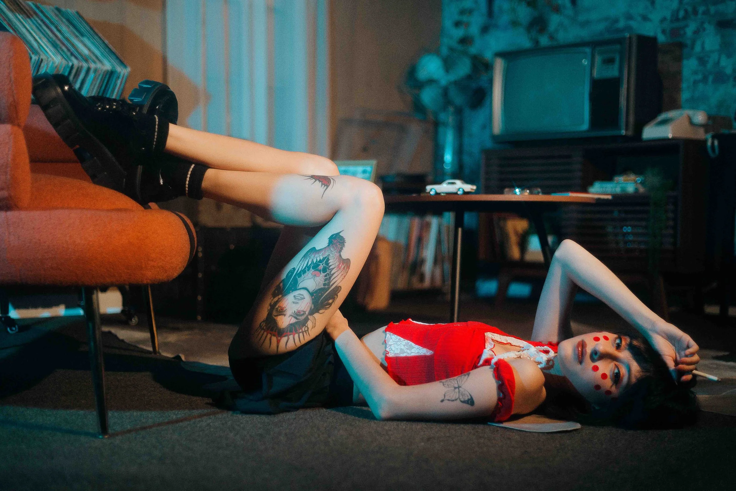 Editorial portrait of a tattooed woman with artistic face paint lying on the floor in a red dress and black shoes, her legs resting on a sofa with one arm stretched above her head