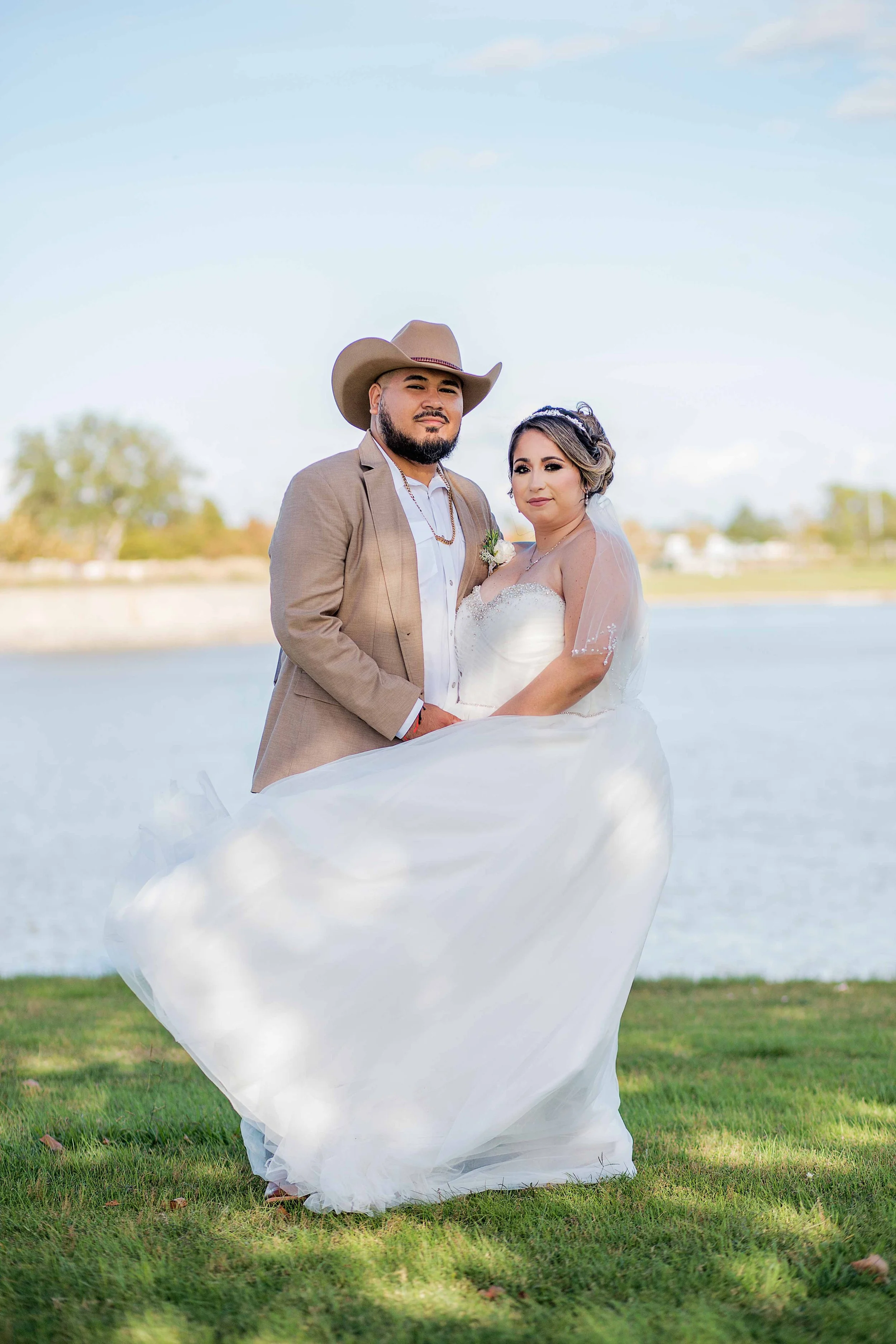 Outdoor wedding portrait in Houston, Texas by Lev’s Photography featuring a newlywed couple standing by a lake, with the groom in a tan suit and cowboy hat and the bride in a white gown and veil embracing