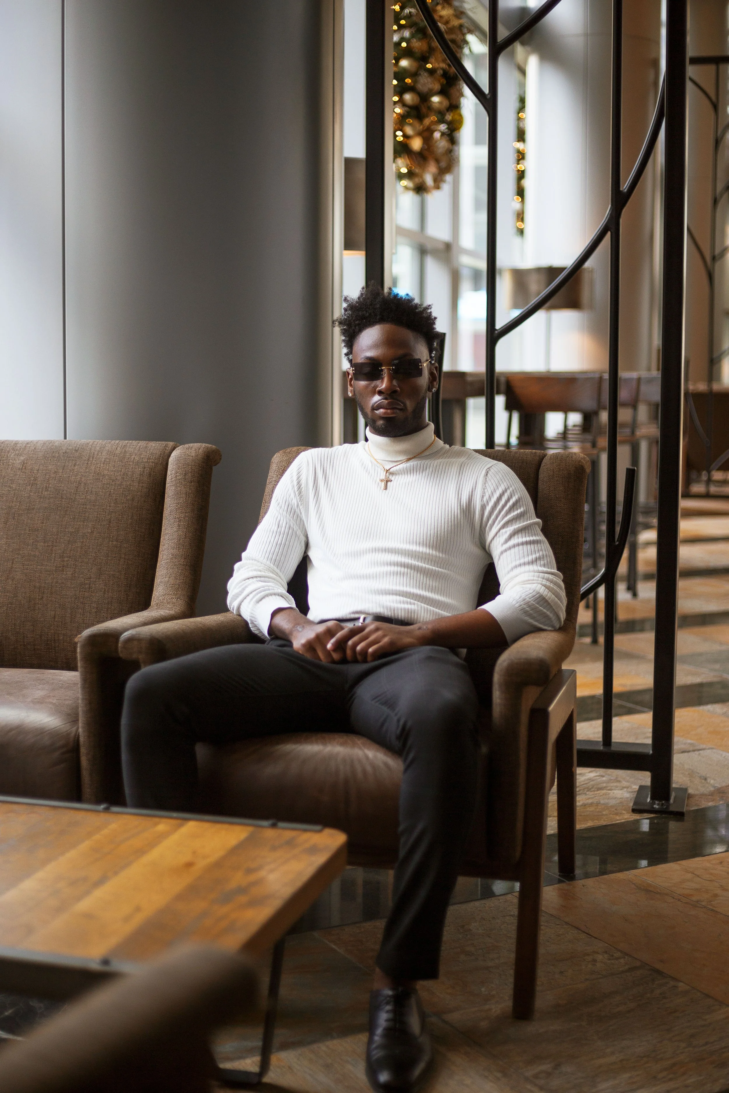 A man seated on a brown armchair in a modern, stylish cafe, wearing sunglasses, a white turtleneck sweater, black pants, and a necklace with a cross, with holiday decorations in the background.