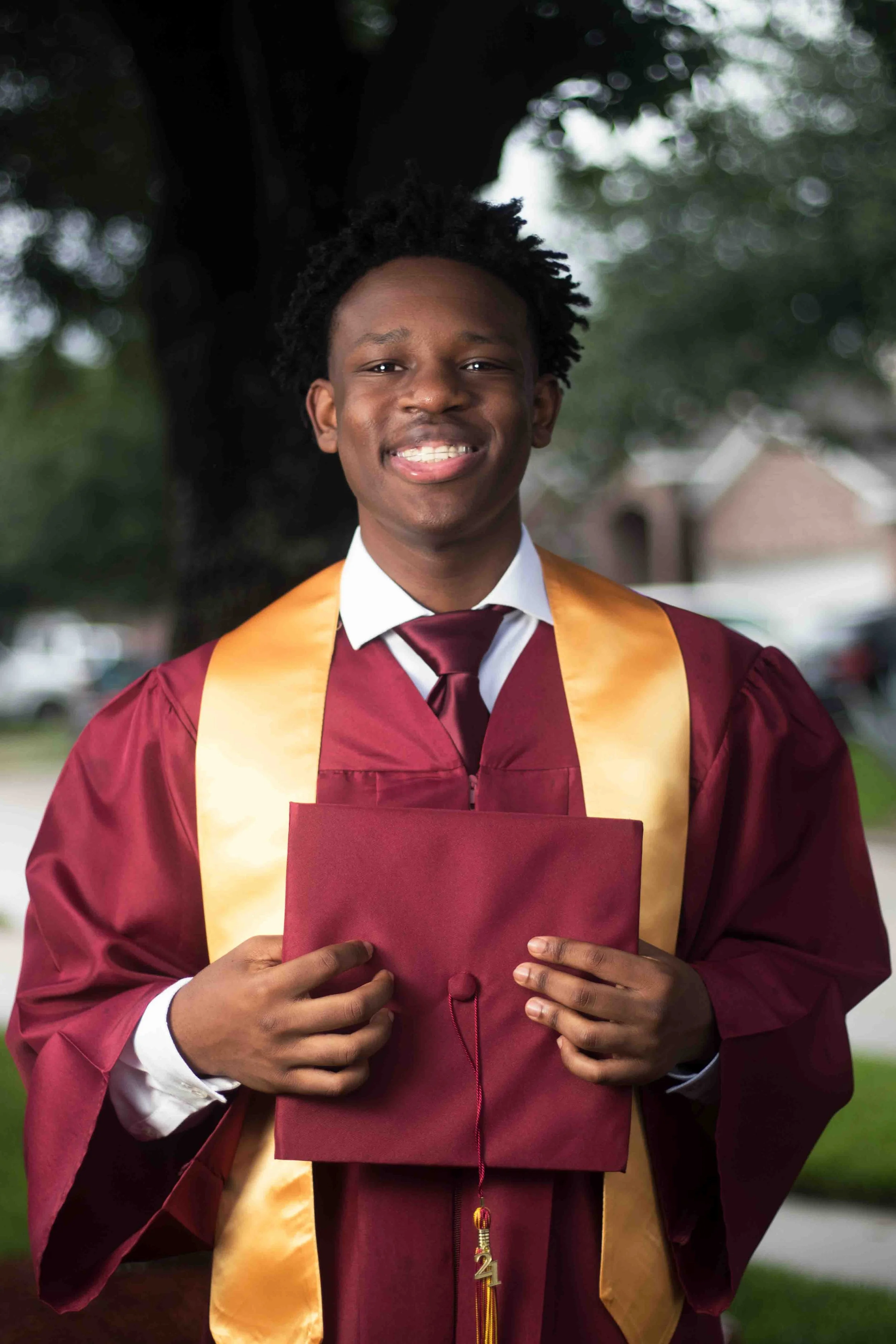Outdoor graduation portrait of a young man in a maroon cap and gown holding his diploma and smiling, standing near trees and houses