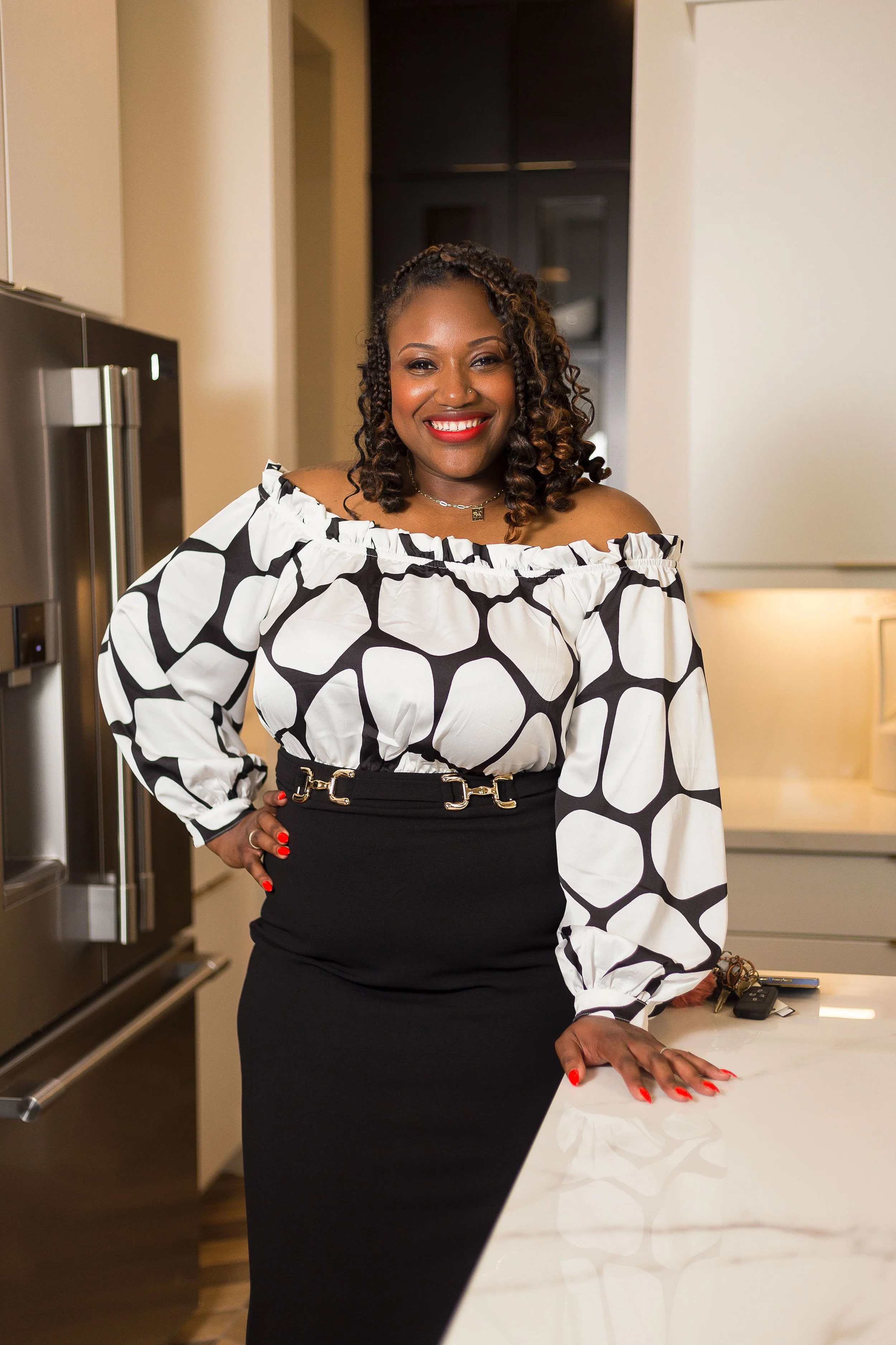 A smiling woman with curly hair wearing a black and white patterned off-shoulder blouse and black high-waisted skirt stands in a modern kitchen, with her left hand resting on a white marble countertop.