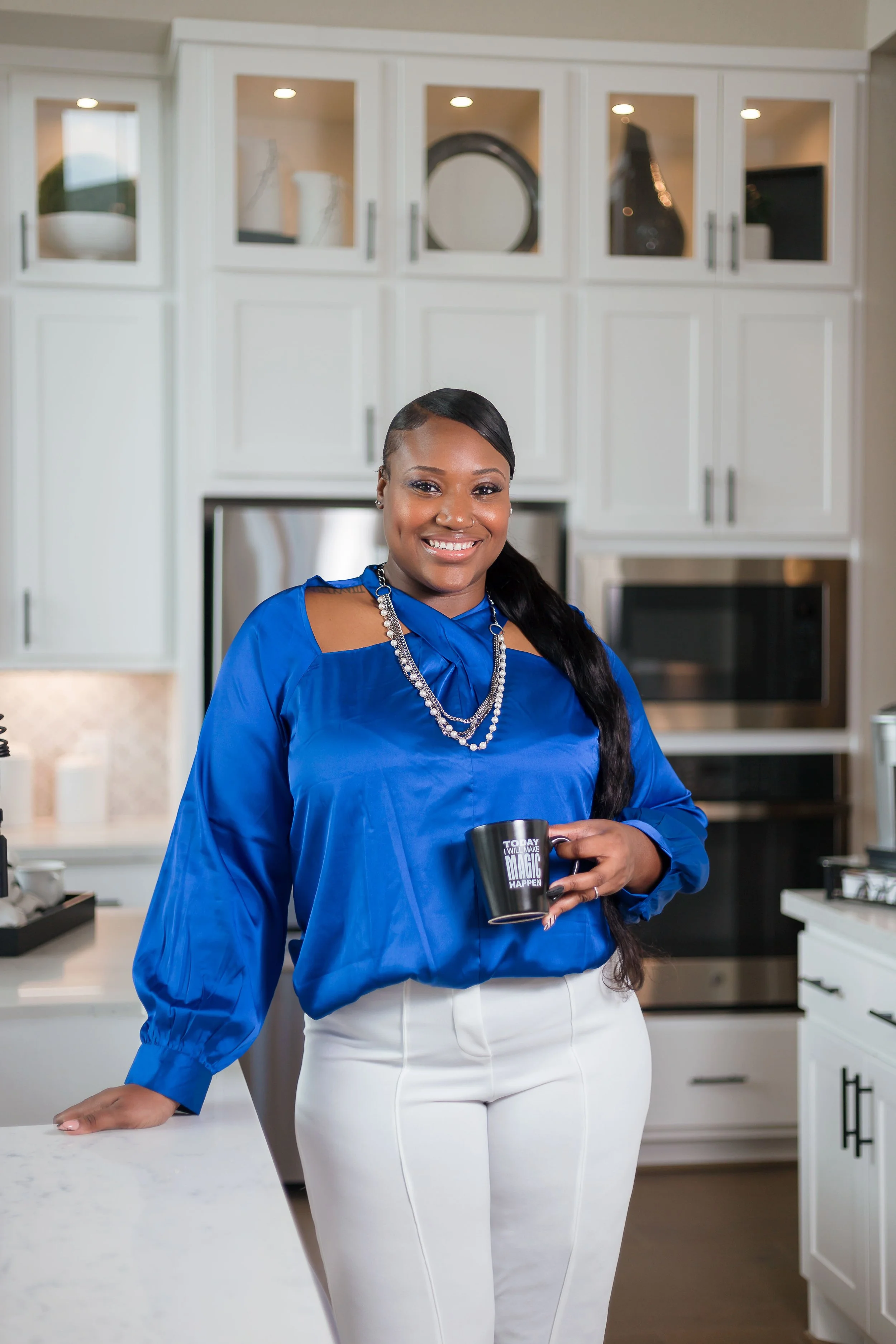 A woman smiling and holding a black mug that says "Today I Will Make Magic Happen" in a modern kitchen with white cabinets.