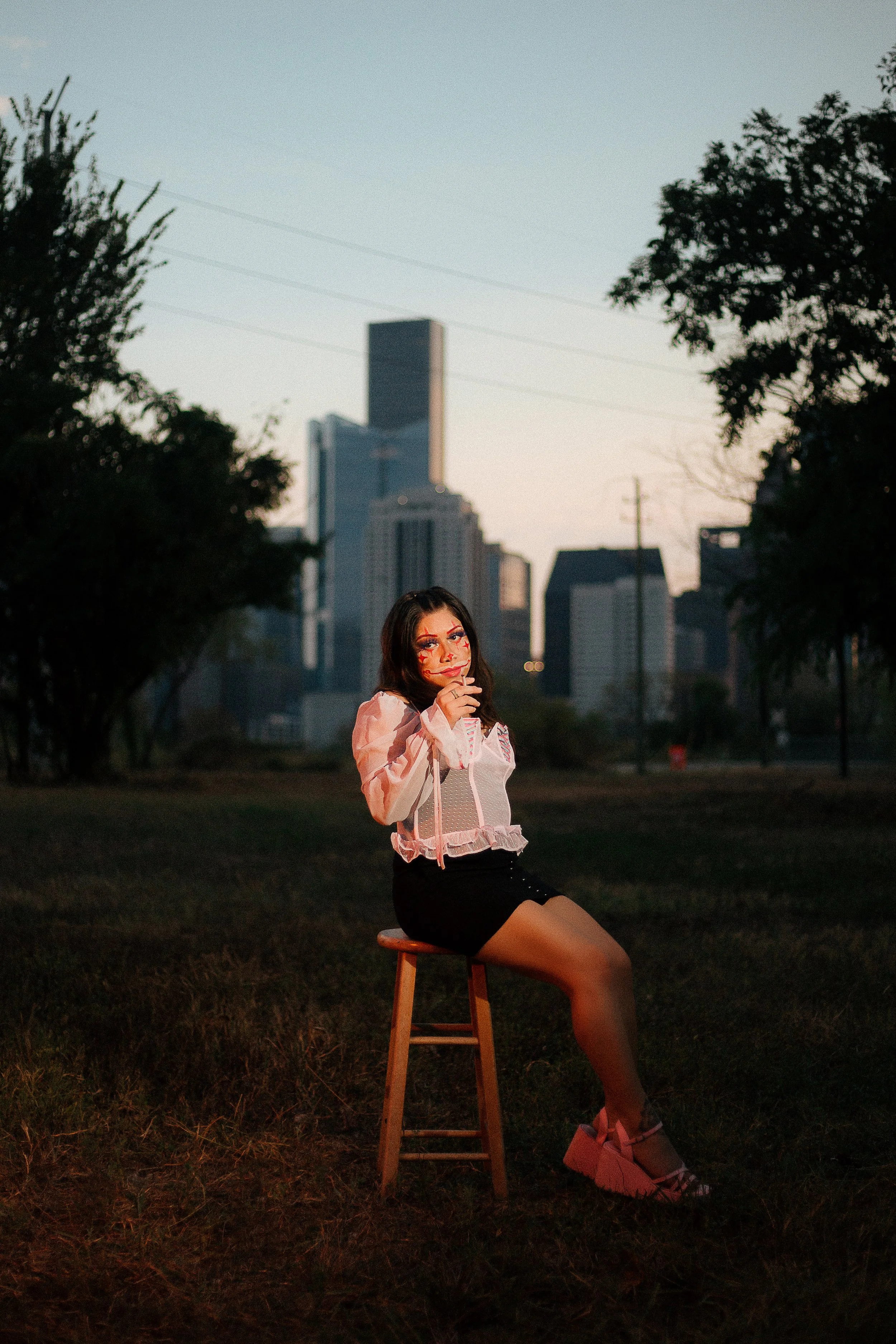 A woman sitting on a wooden stool in a park at dusk, with a city skyline in the background. She has dark hair, wears a light pink jacket, black skirt, and pink platform shoes. She has makeup with red accents around her eyes and face.