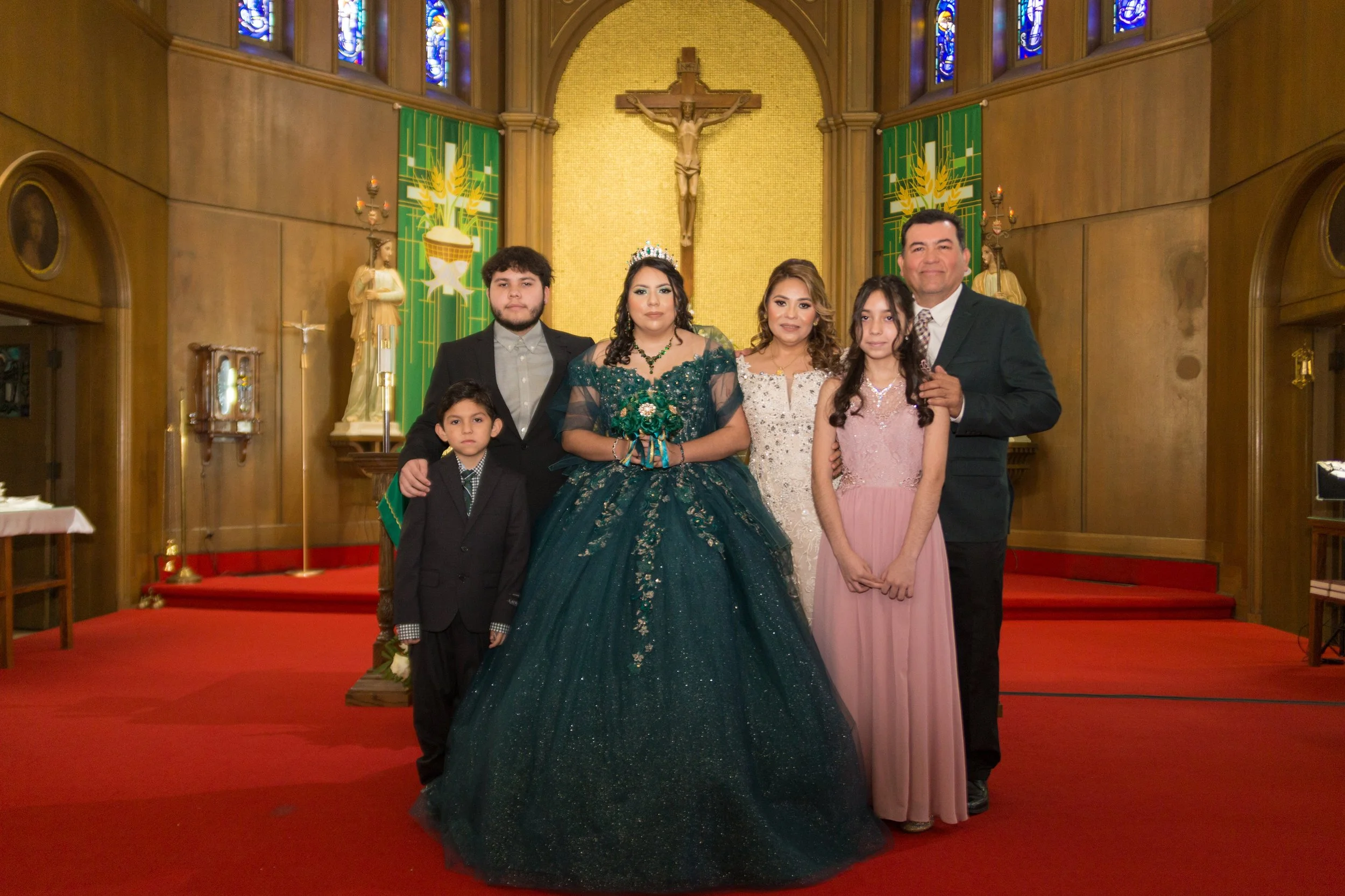 Family in formal attire posing inside a church, with a woman in an elaborate green gown and a tiara, surrounded by children and adults, in front of a crucifix and religious decorations.