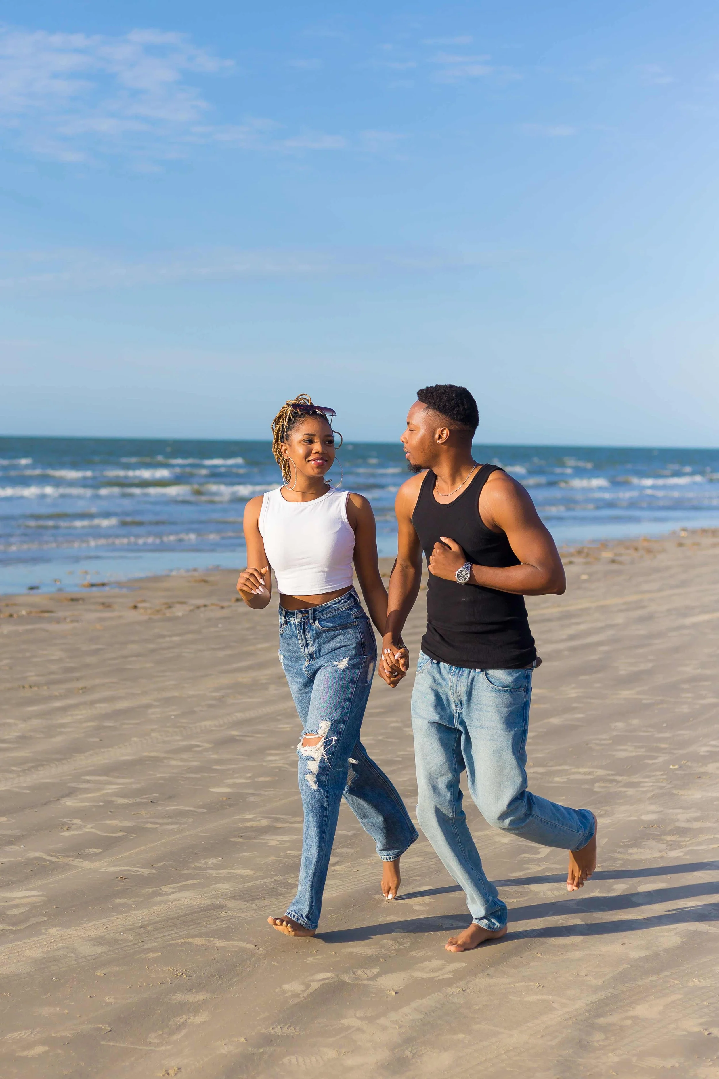 Beach couple portrait in Galveston, Texas by Lev’s Photography featuring a young couple holding hands and walking barefoot along the shoreline with the ocean and blue sky in the background