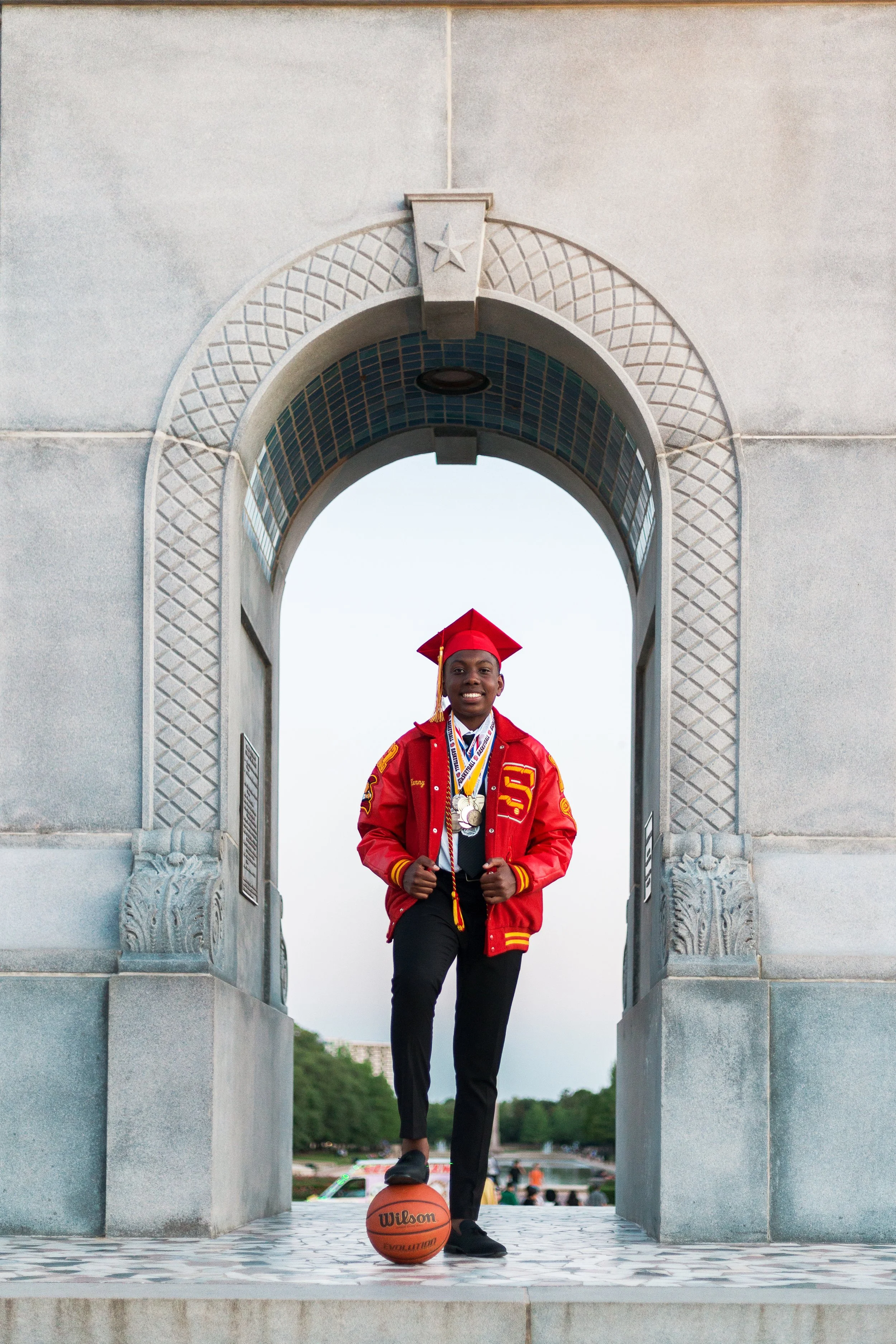 A young man in a red graduation cap and gown with multiple medals, standing on a pedestal with a basketball, in front of a large stone archway.
