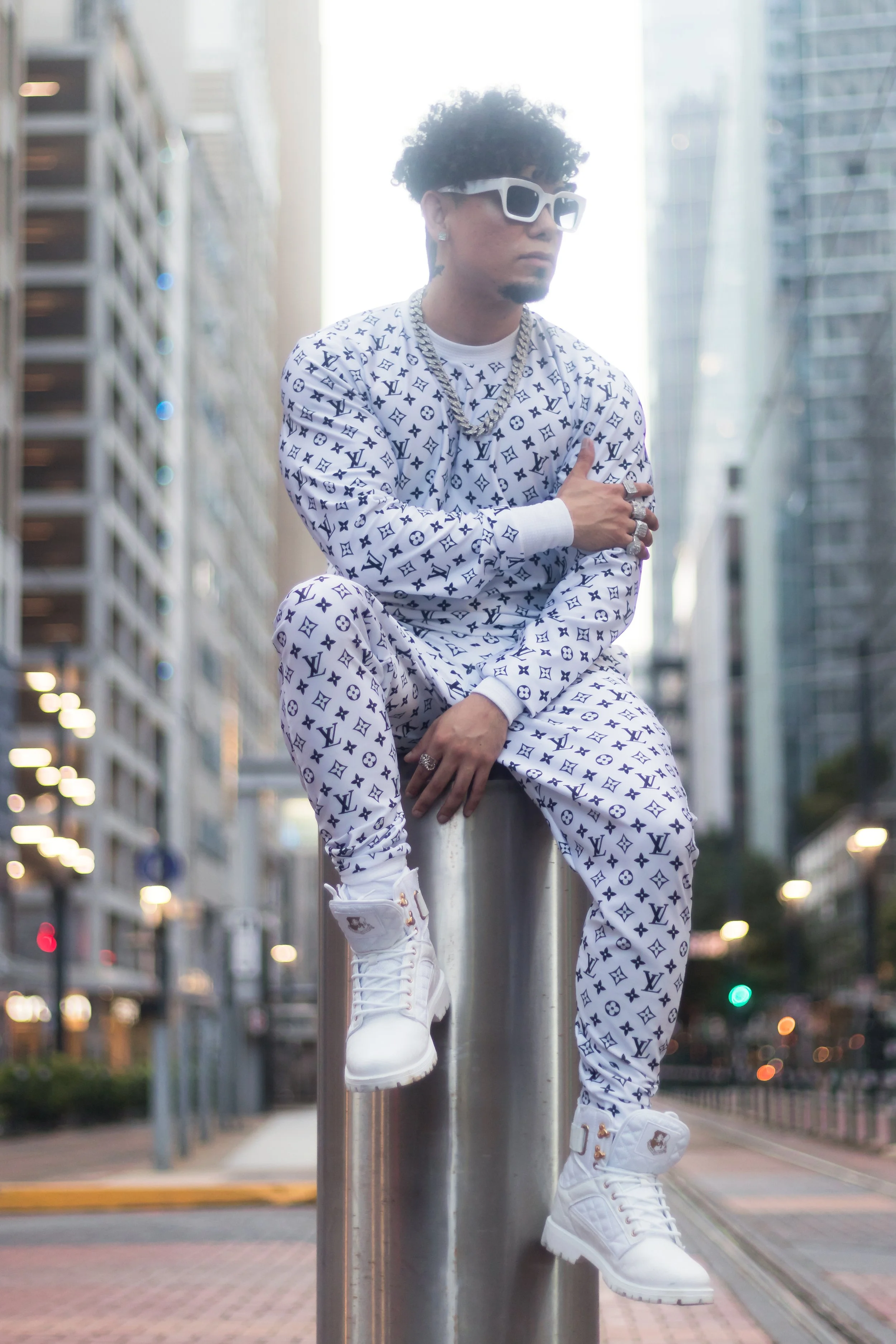 A man dressed in white Louis Vuitton matching sweatshirt and pants with black logos, wearing white sunglasses, jewelry, and white high-top sneakers, sitting on a metal post in an urban city street with tall modern buildings in the background.