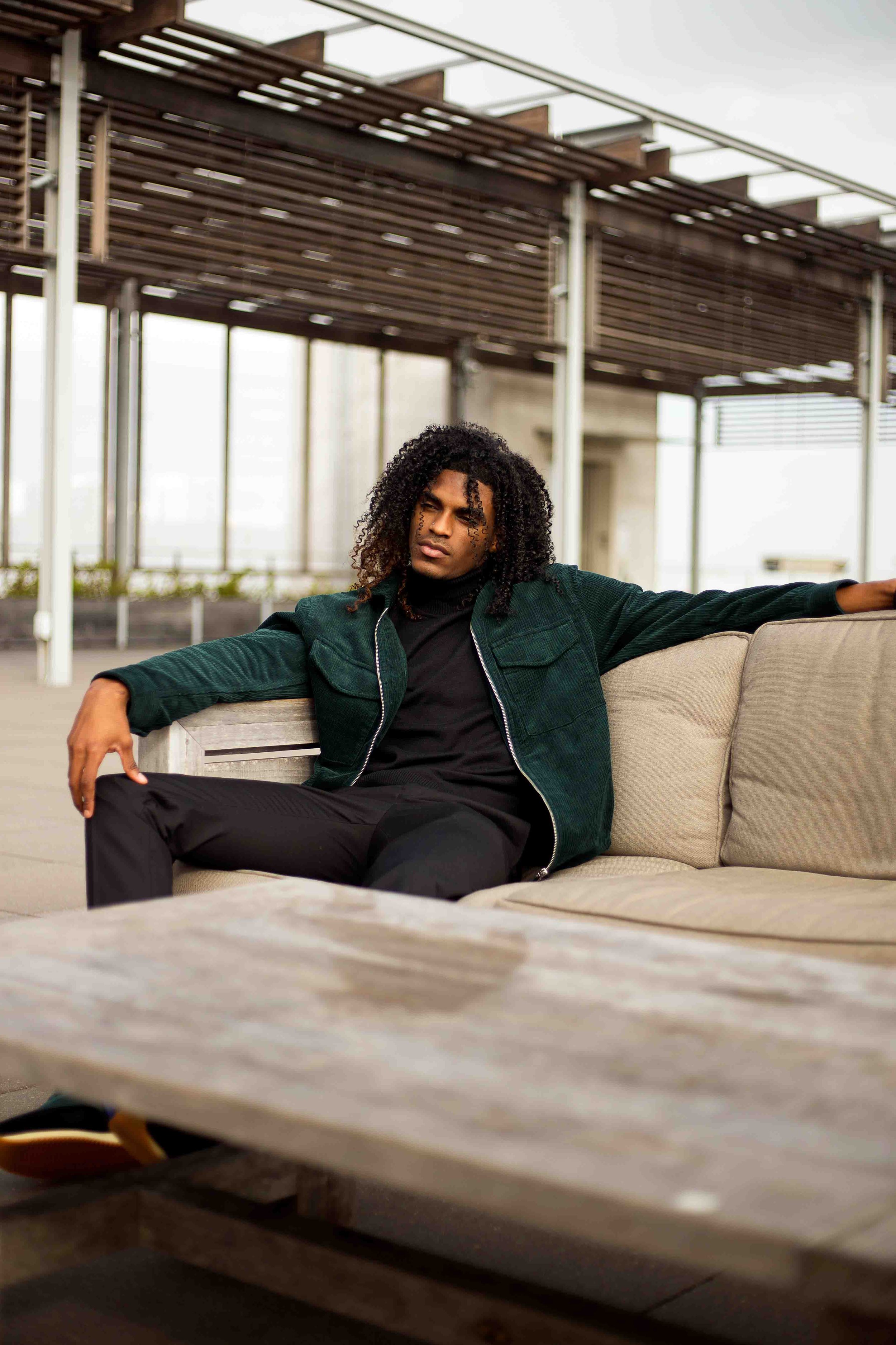 Outdoor lifestyle portrait in Houston, Texas by Lev’s Photography featuring a man with curly hair sitting on a beige sofa wearing a dark green jacket and black pants with a modern building in the background