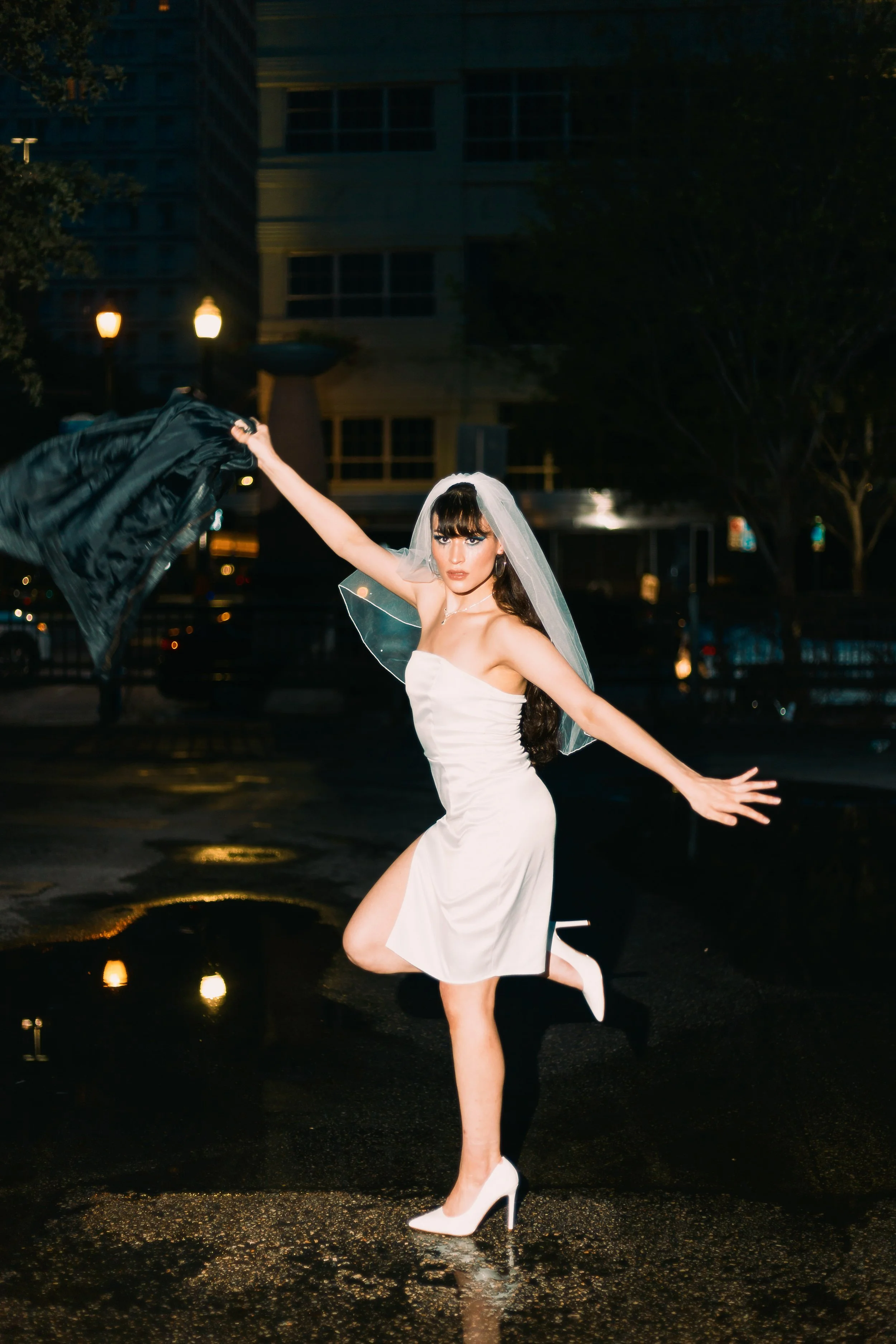A woman in a white dress and heels, wearing a wedding veil, dancing outside at night on a wet pavement, with a cityscape in the background.