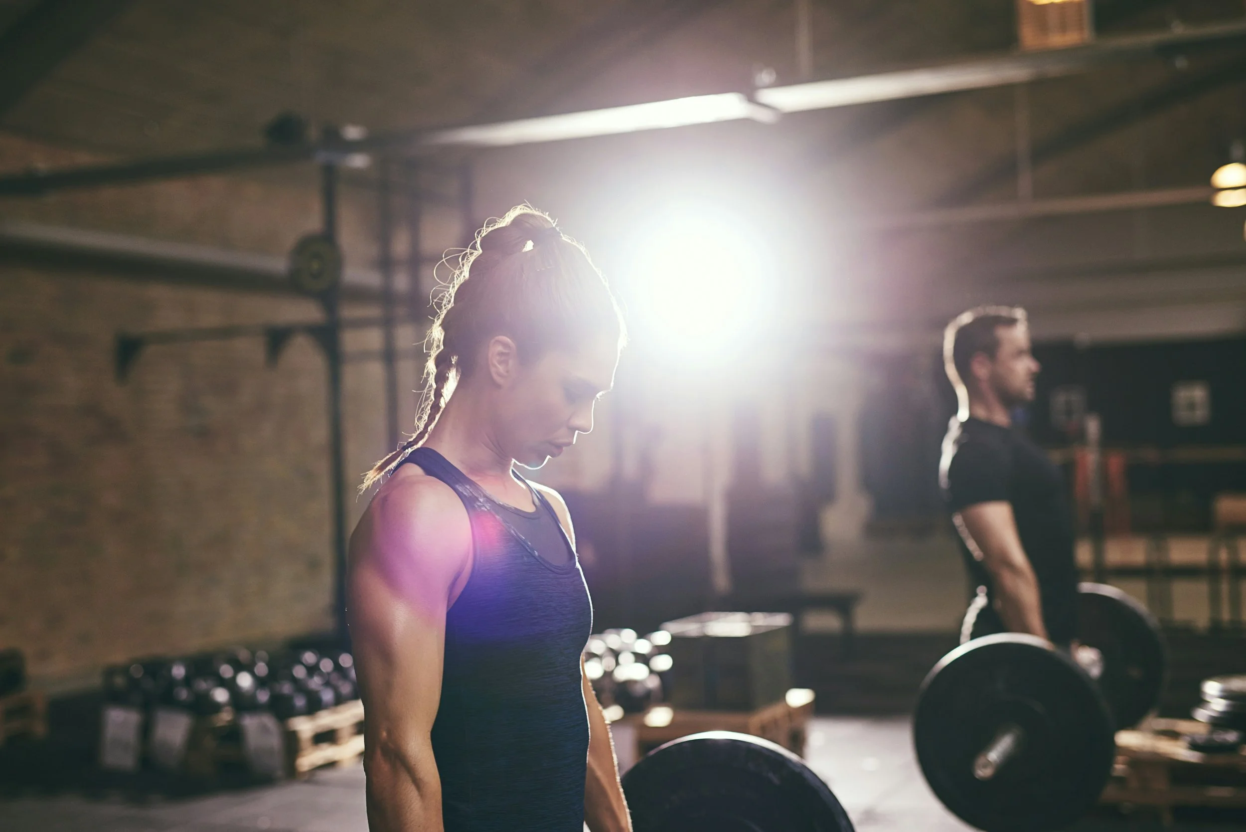 A woman and a man lifting dumbbells in a gym with exposed brick walls, illuminated by bright overhead lighting.