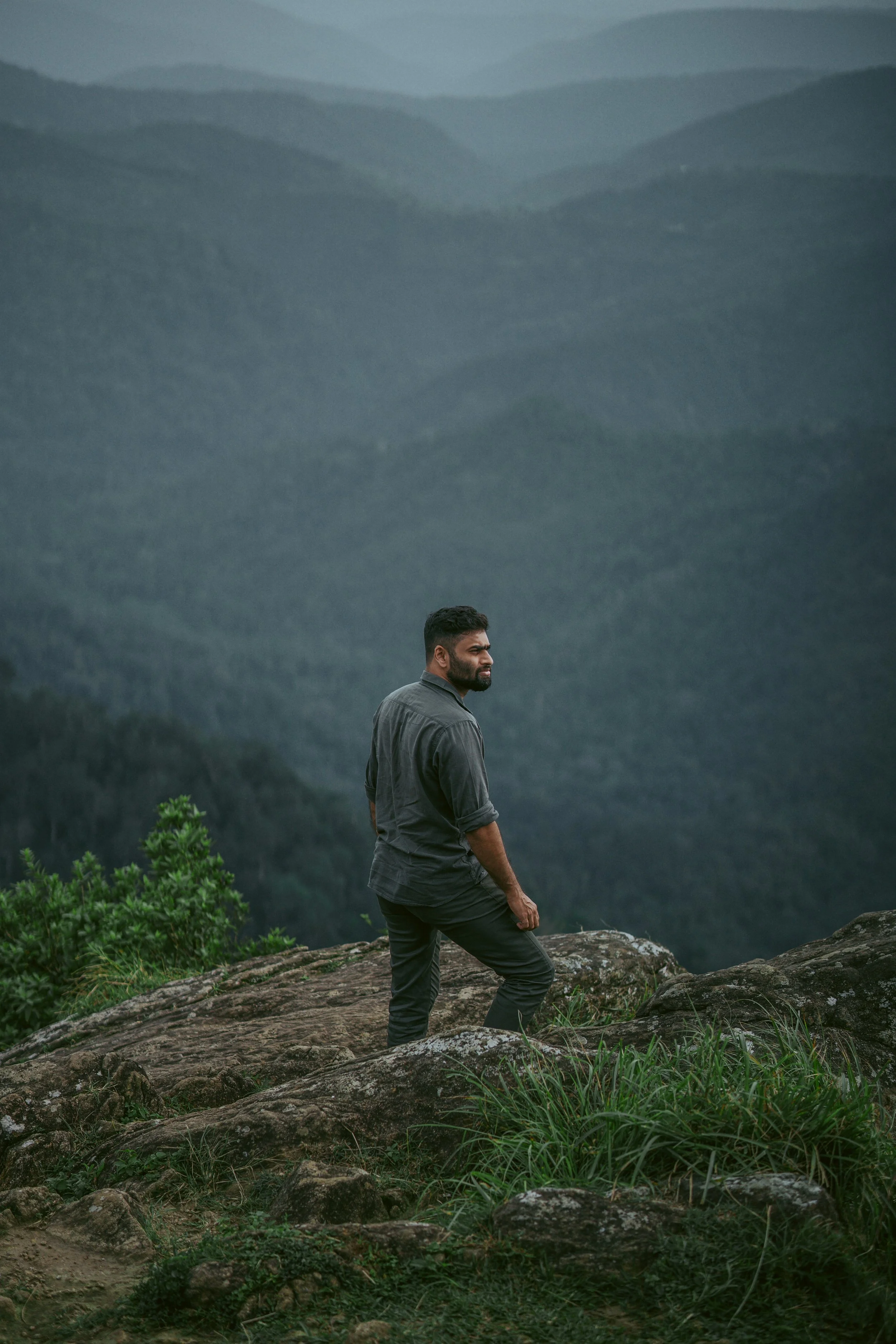 A man with dark hair and a beard standing on rocky terrain with green plants, looking to his left, mountain landscape in the background under a cloudy sky.