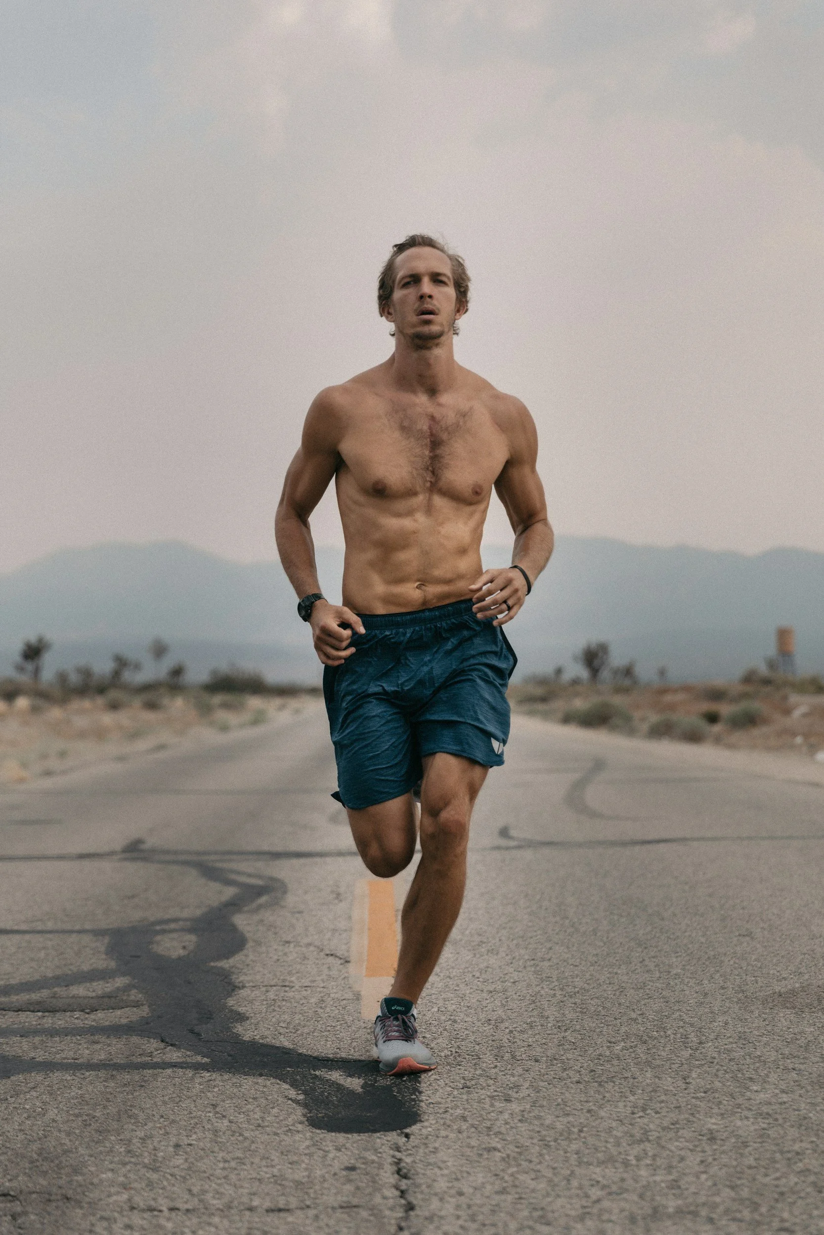 A man runs shirtless on an empty road with mountain scenery in the background.