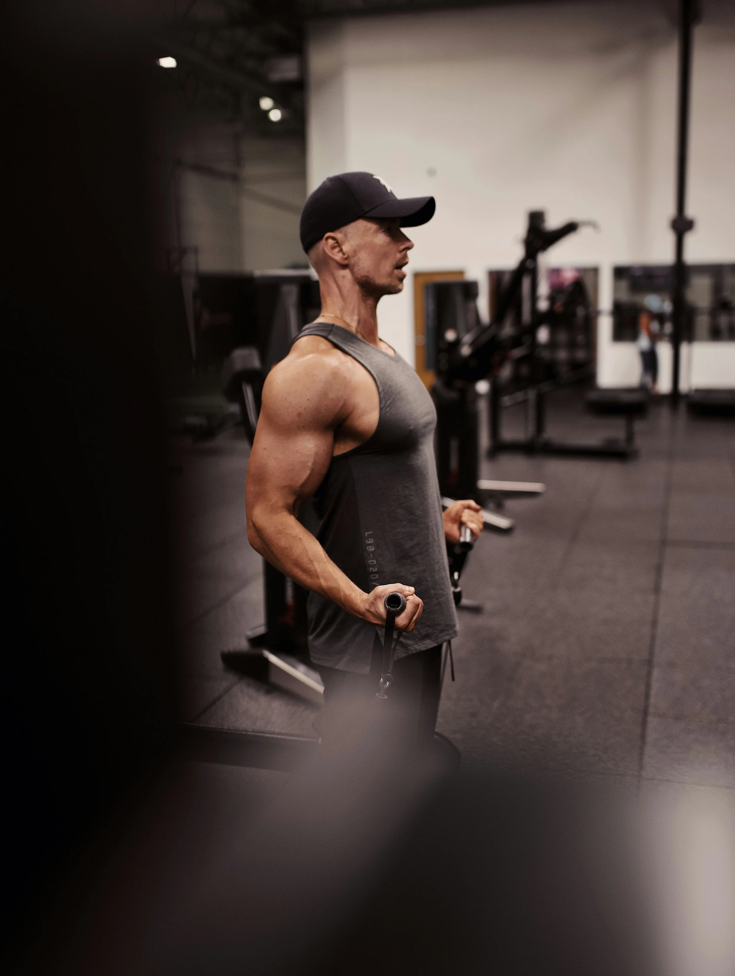 A man with a black baseball cap, gray tank top, and short hair using a jump rope in a gym. The gym has various workout equipment and machines in the background.