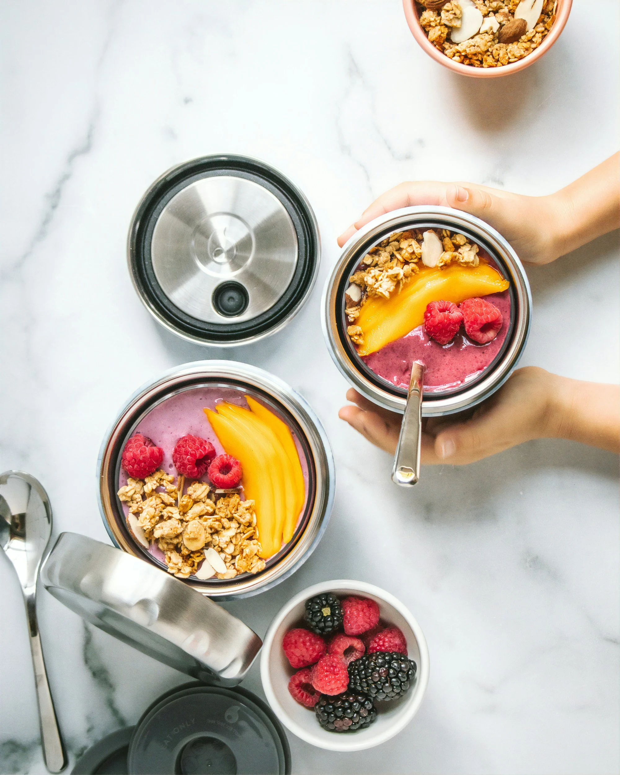 Two bowls of mixed berry smoothies topped with granola, sliced peaches, raspberries, and almond slices on a white marble surface, with a small bowl of blackberries and raspberries nearby.